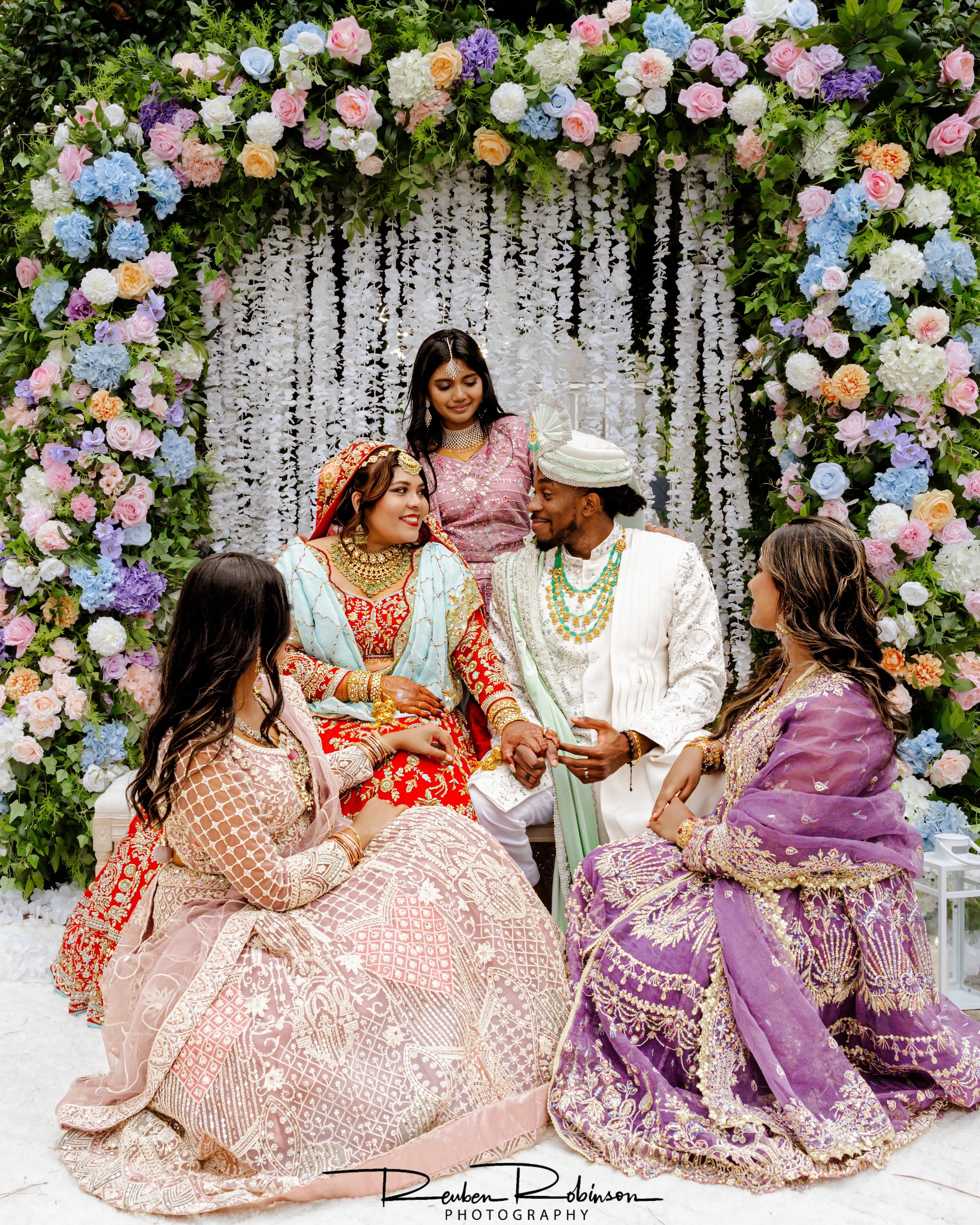 A bride and groom seated under floral arch at wedding ceremony with bridesmaids and friends around them.