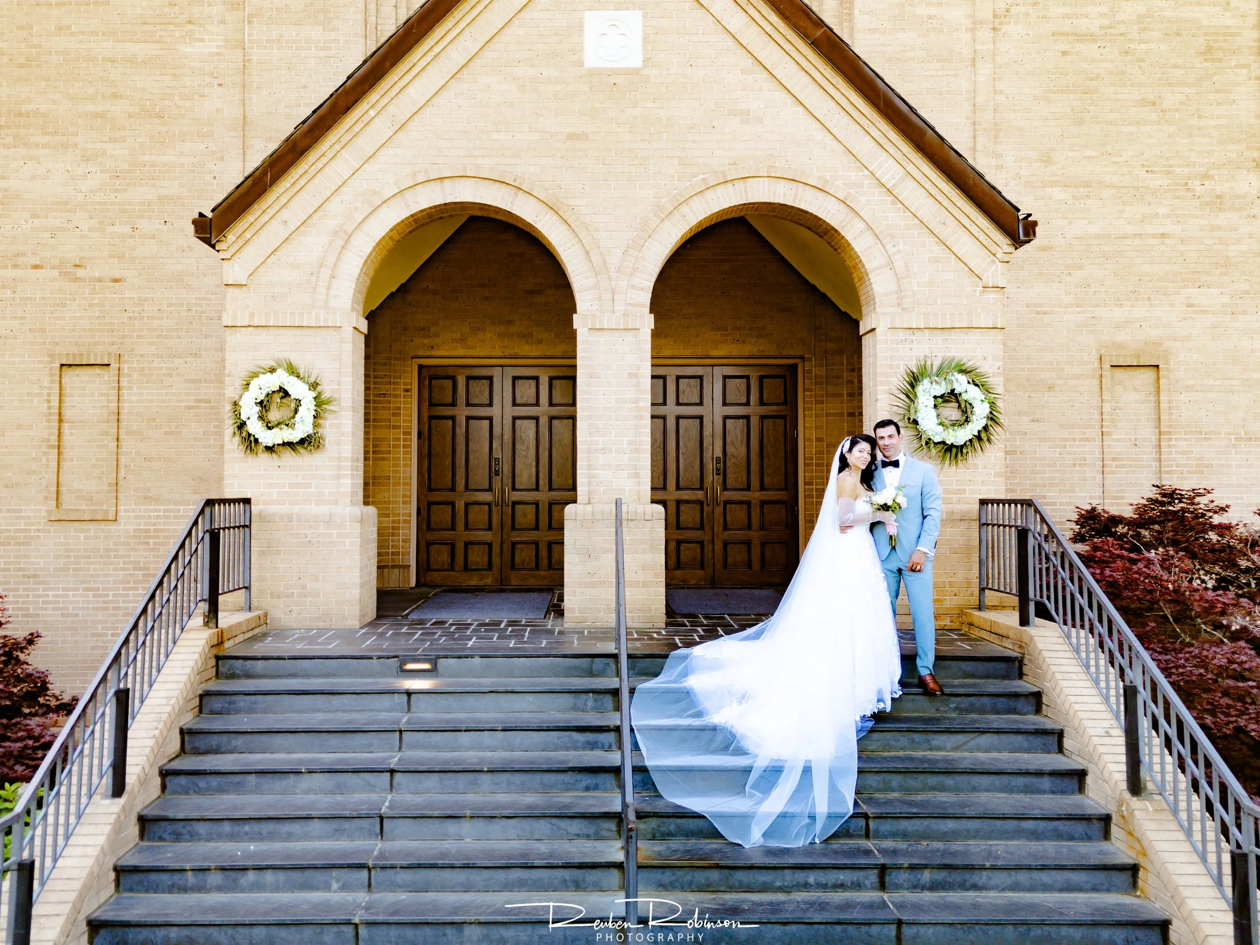 A bride and groom standing on the steps of a church, with two floral wreaths on the brick wall behind them.