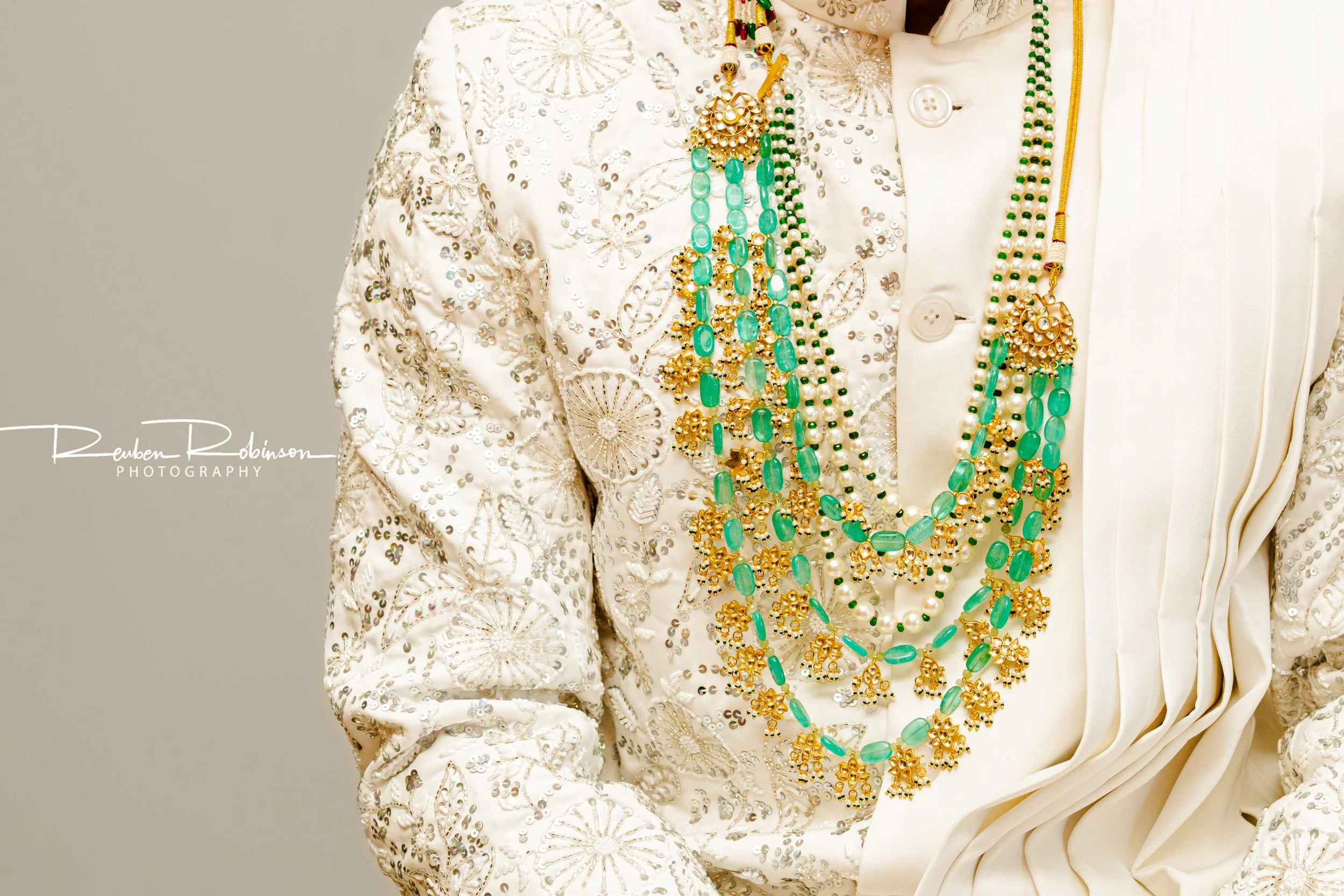 Close-up of a person wearing a white embroidered jacket adorned with sequins, layered with multiple strands of turquoise, pearl, and gold jewelry, including necklaces with beads and intricate gold designs.