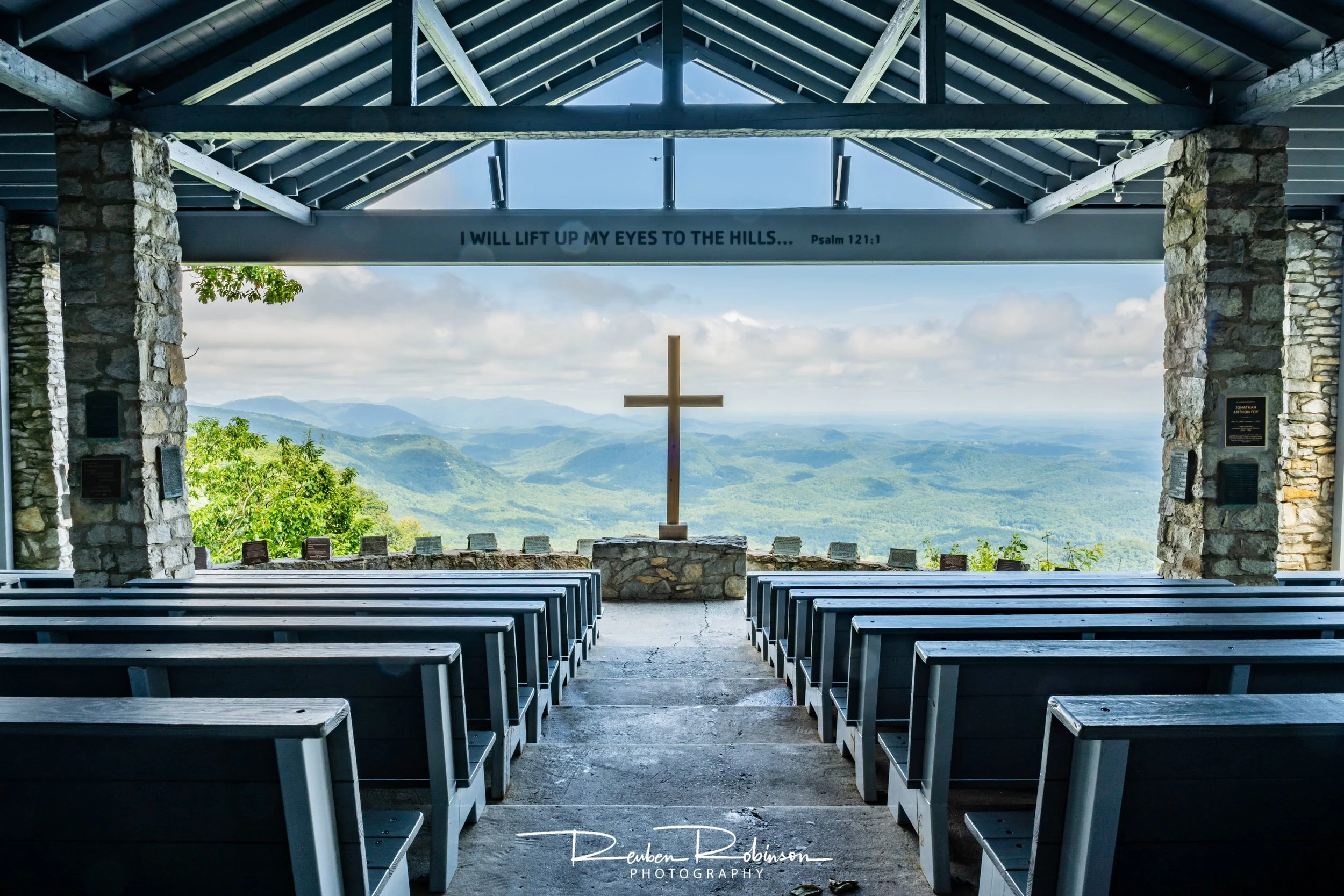 Interior of a scenic outdoor chapel with wooden benches, stone pillars, and a wooden cross, overlooking a panoramic view of green mountains and blue sky.