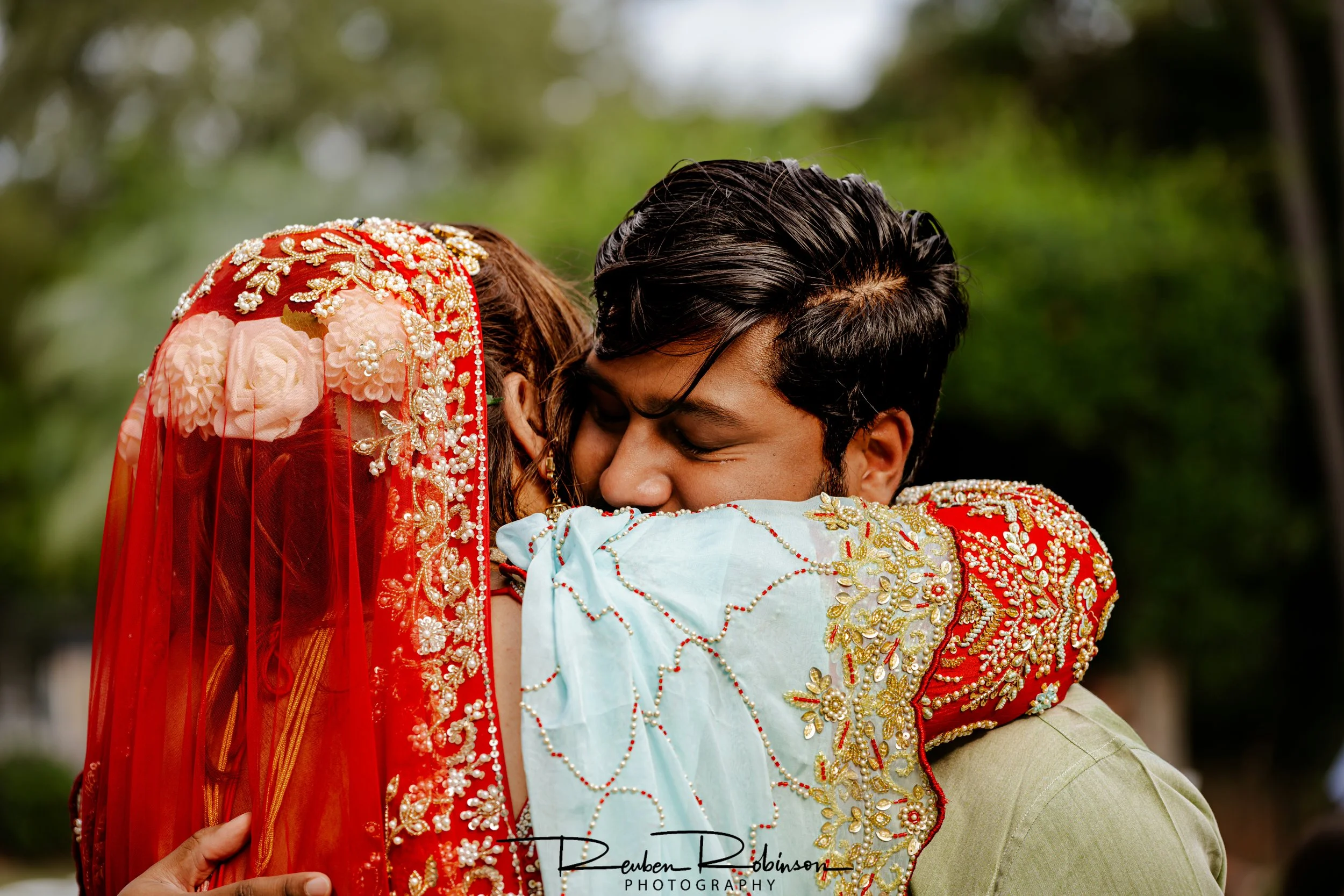 A couple dressed in traditional Indian wedding attire embracing each other outdoors. The woman wears a red lehenga with gold embroidery and a red veil with floral embellishments, while the man wears a light-colored sherwani with gold embroidery.