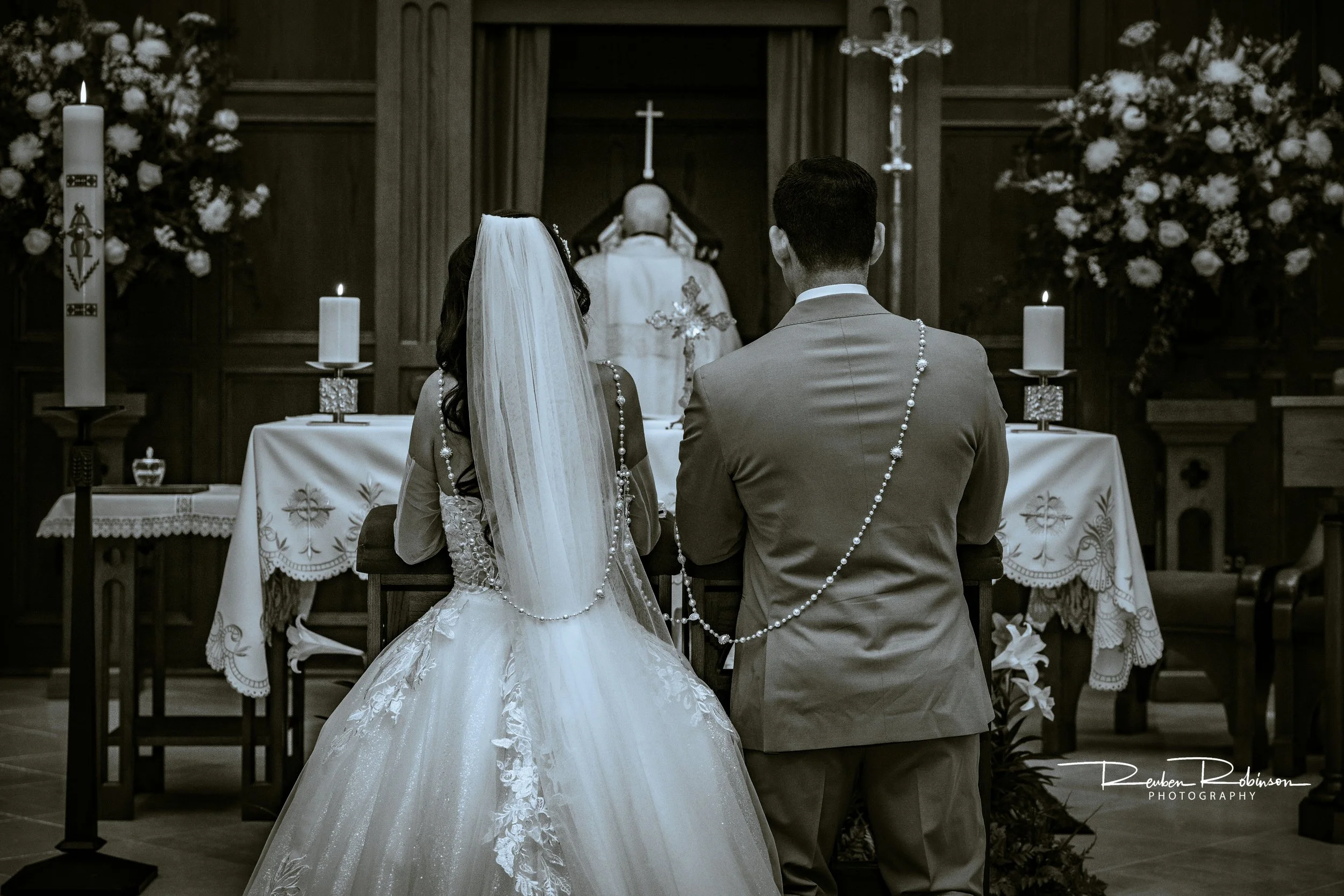 A bride and groom kneeling at an altar during a wedding ceremony inside a church. The bride is wearing a white wedding gown with lace details and a veil, and the groom is in a light-colored suit. They are facing a priest, with candles and floral arra