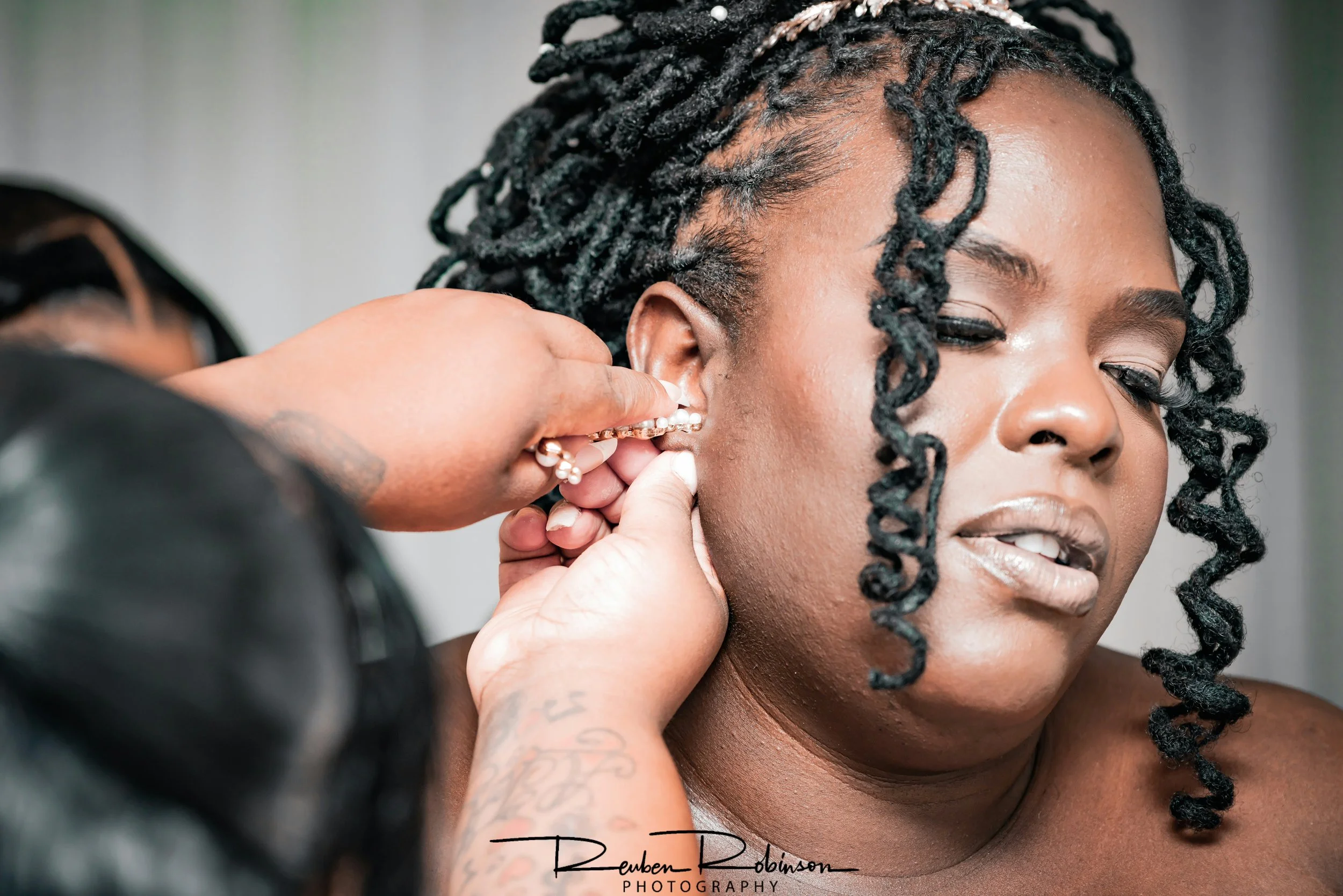 A woman with styled dreadlocks getting her ear pierced by a person wearing earrings, with a focus on the woman's face and the piercing process.