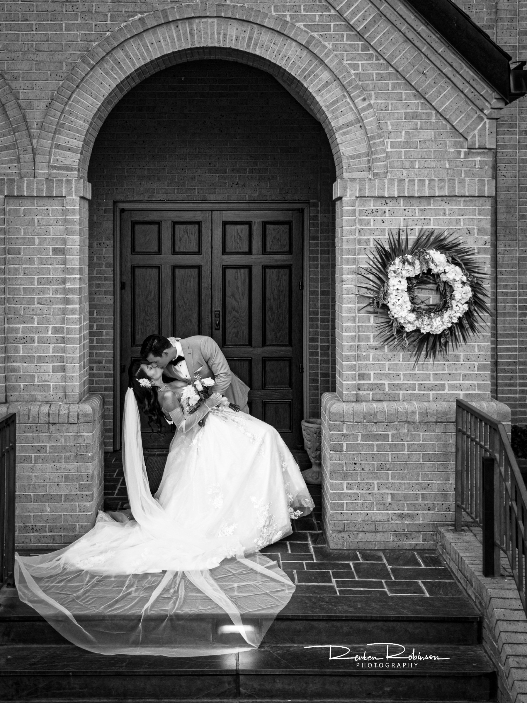 A bride and groom share a kiss in front of a brick archway doorway, with a floral wreath hanging on the wall to the right, in black and white photography.