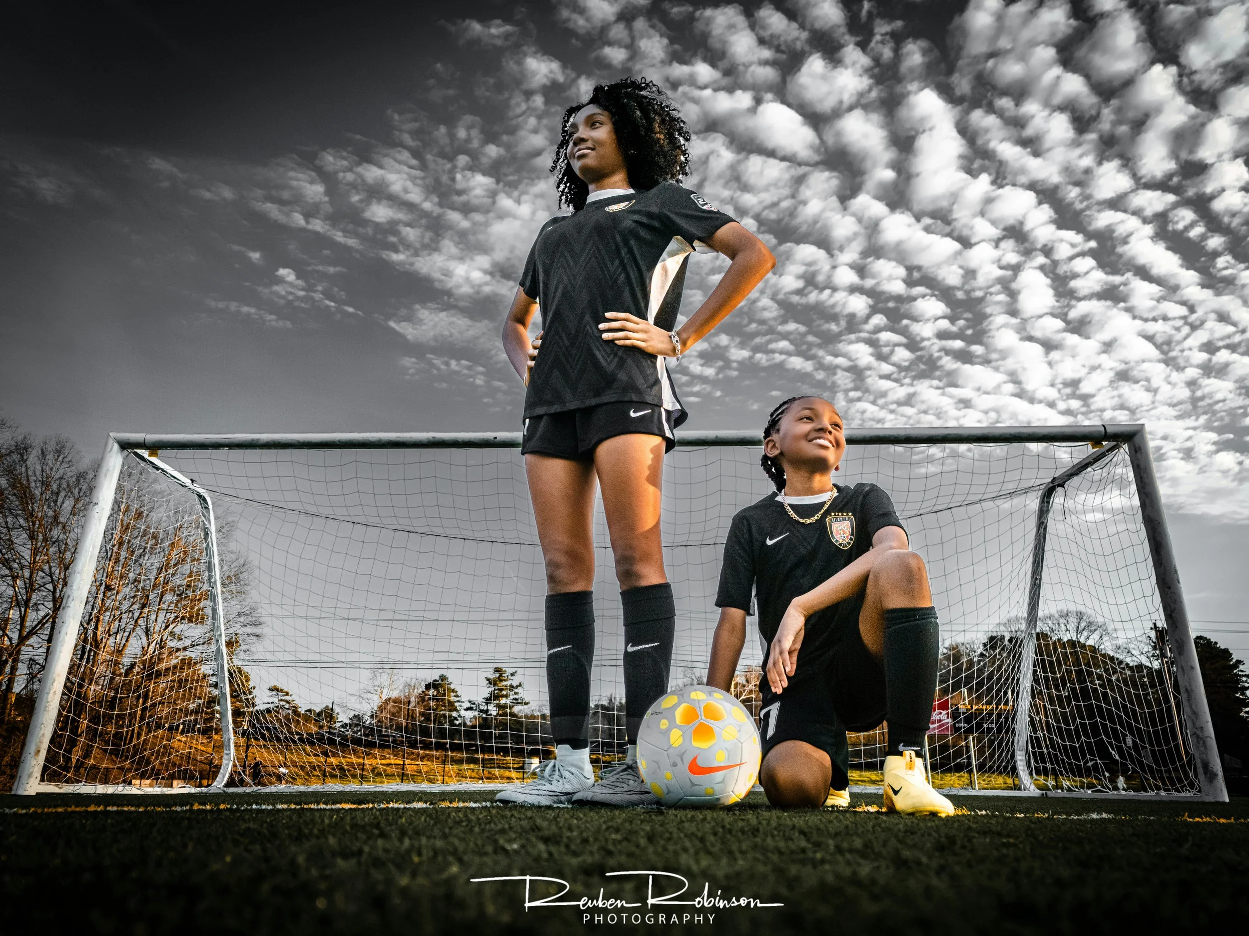 Two female soccer players in black uniforms on a soccer field, one standing and the other kneeling next to a soccer ball, with a goalpost behind them and a cloudy sky overhead.