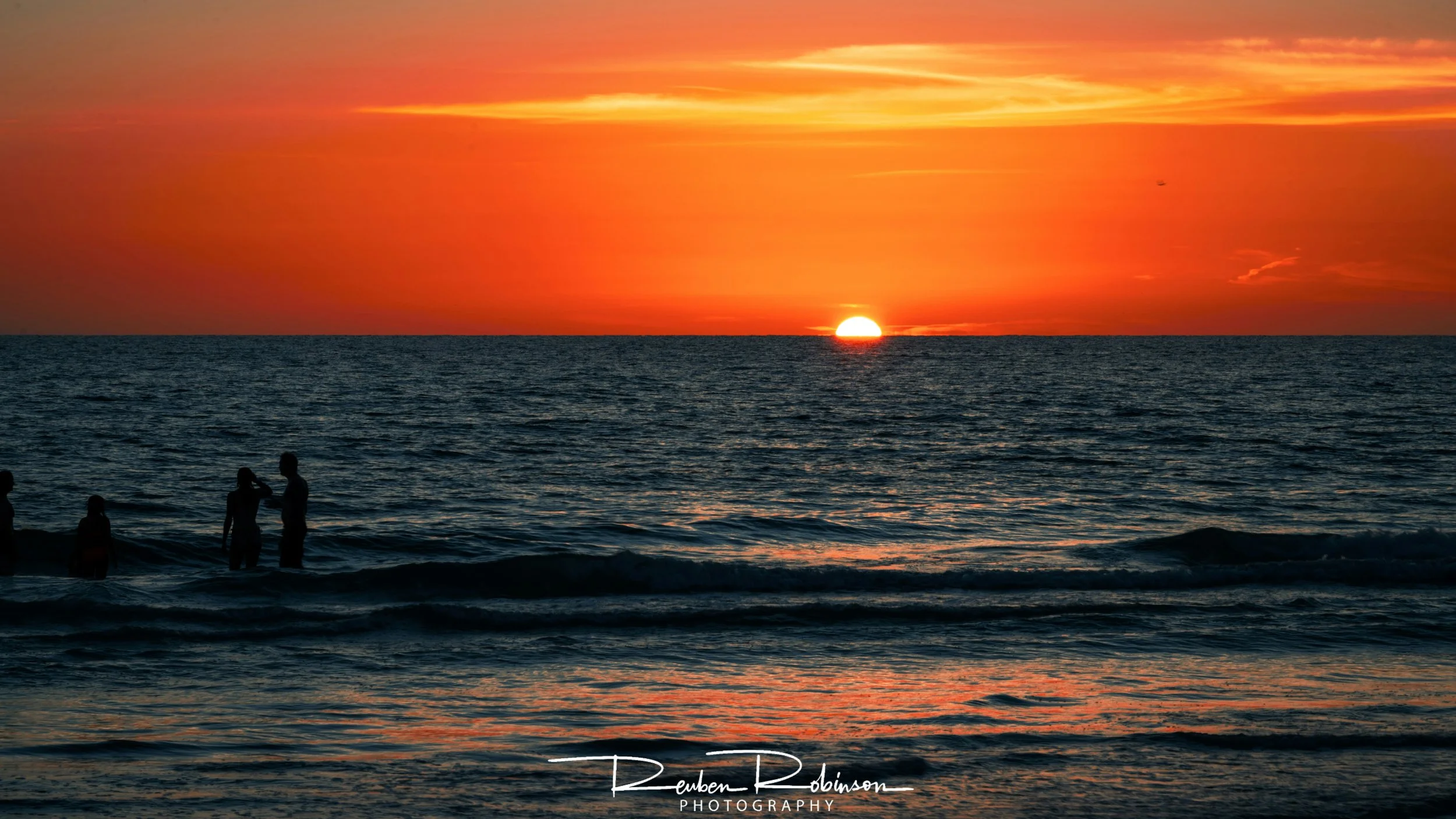 Sun setting on the ocean horizon with a vibrant orange and yellow sky, and silhouettes of people in the water near the shore.