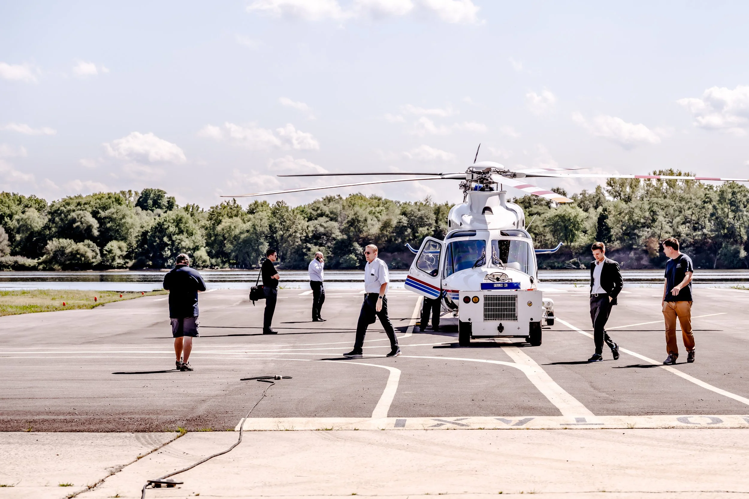 A helicopter on the tarmac with people around it, some in business attire, others in casual clothes, near a body of water and trees under a partly cloudy sky.