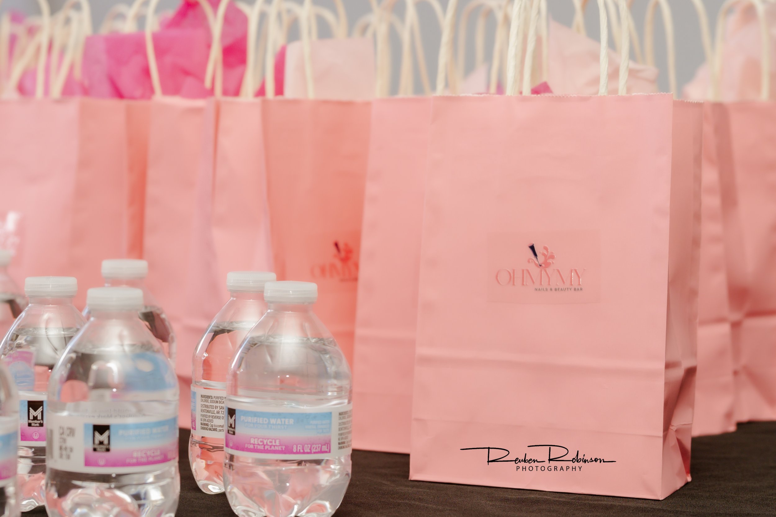 Pink gift bags with a logo and text, and clear bottled water on a table.