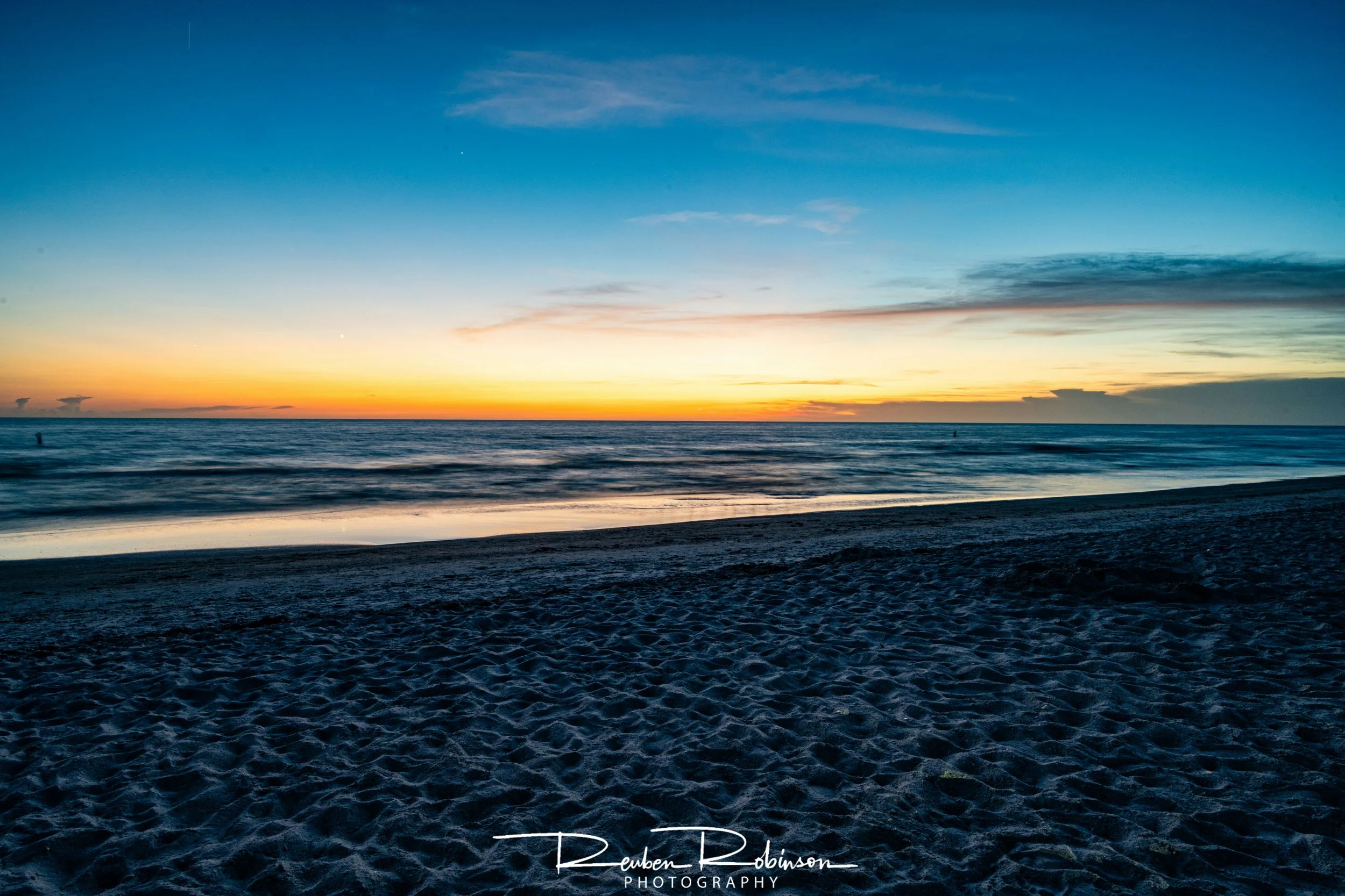 A serene beach scene during sunset with a colorful sky, calm ocean, and sandy shore in the foreground.