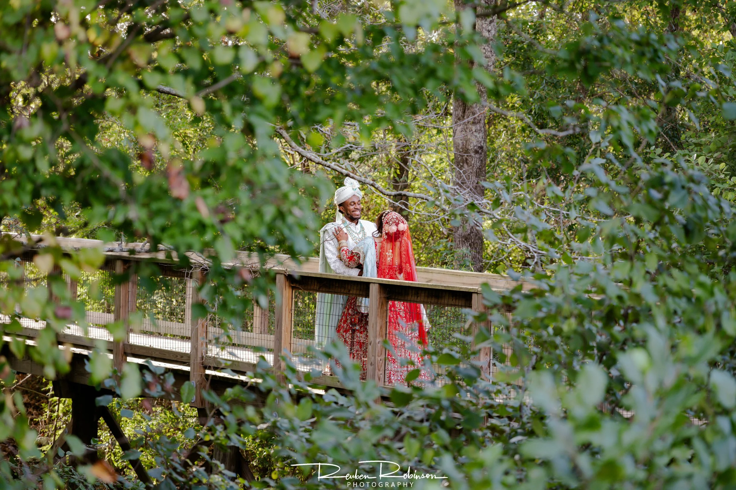 A couple dressed in traditional Indian wedding attire standing on a wooden bridge surrounded by lush green trees.