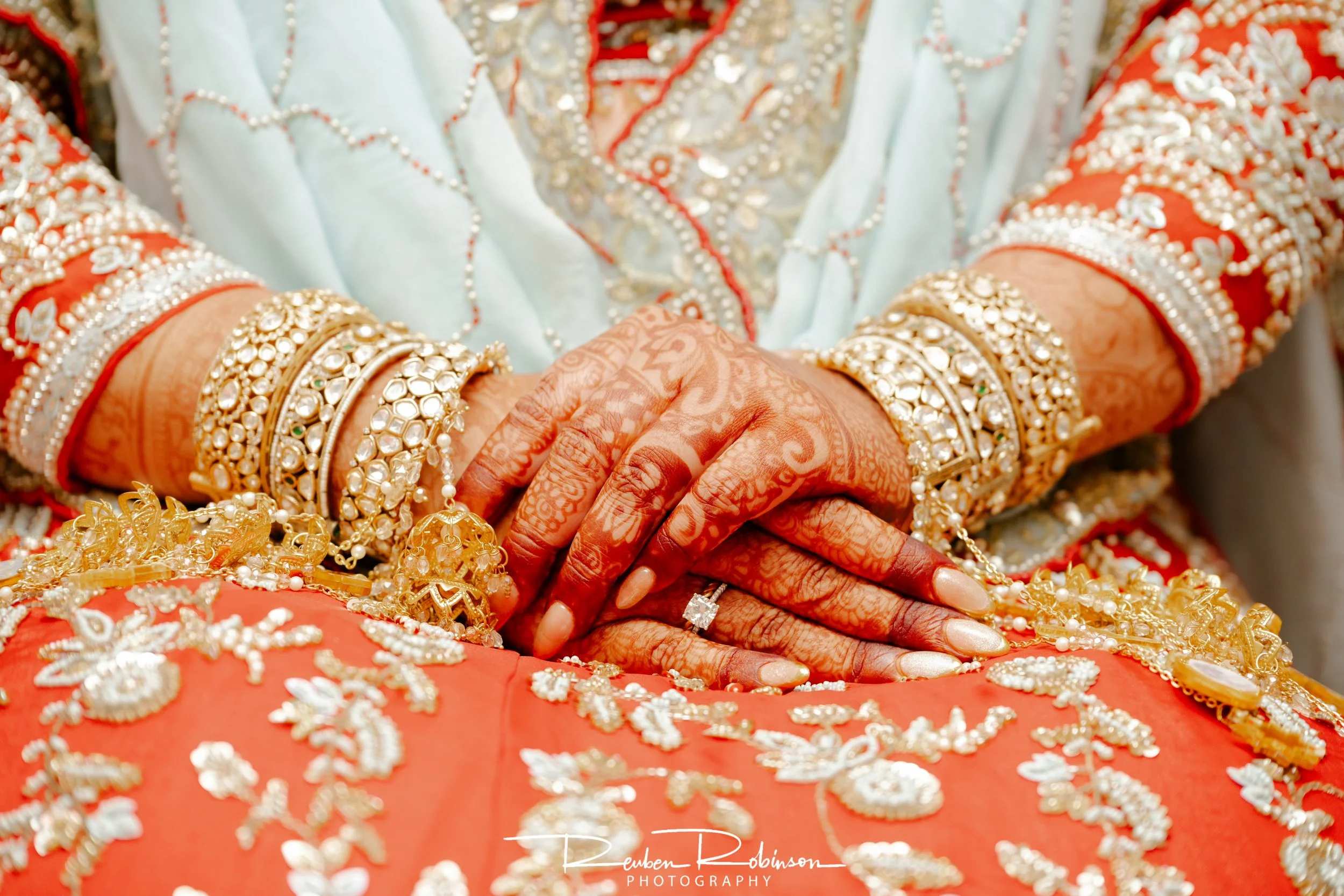 Close-up of a woman wearing traditional Indian wedding attire with intricate gold jewelry, bangles, and henna designs on her hands, sitting against an ornate, embroidered red and gold fabric background.