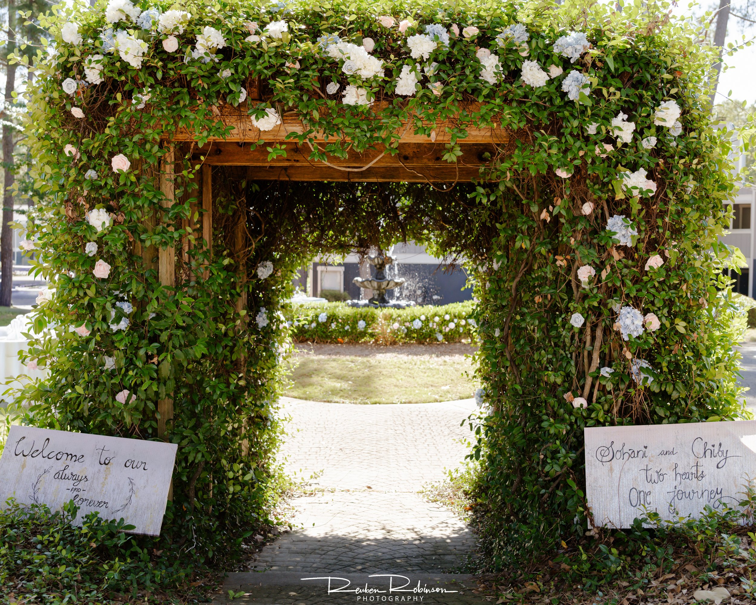 A decorative archway covered with white and pale pink roses and green foliage, leading to a garden area with a fountain and a paved pathway.
