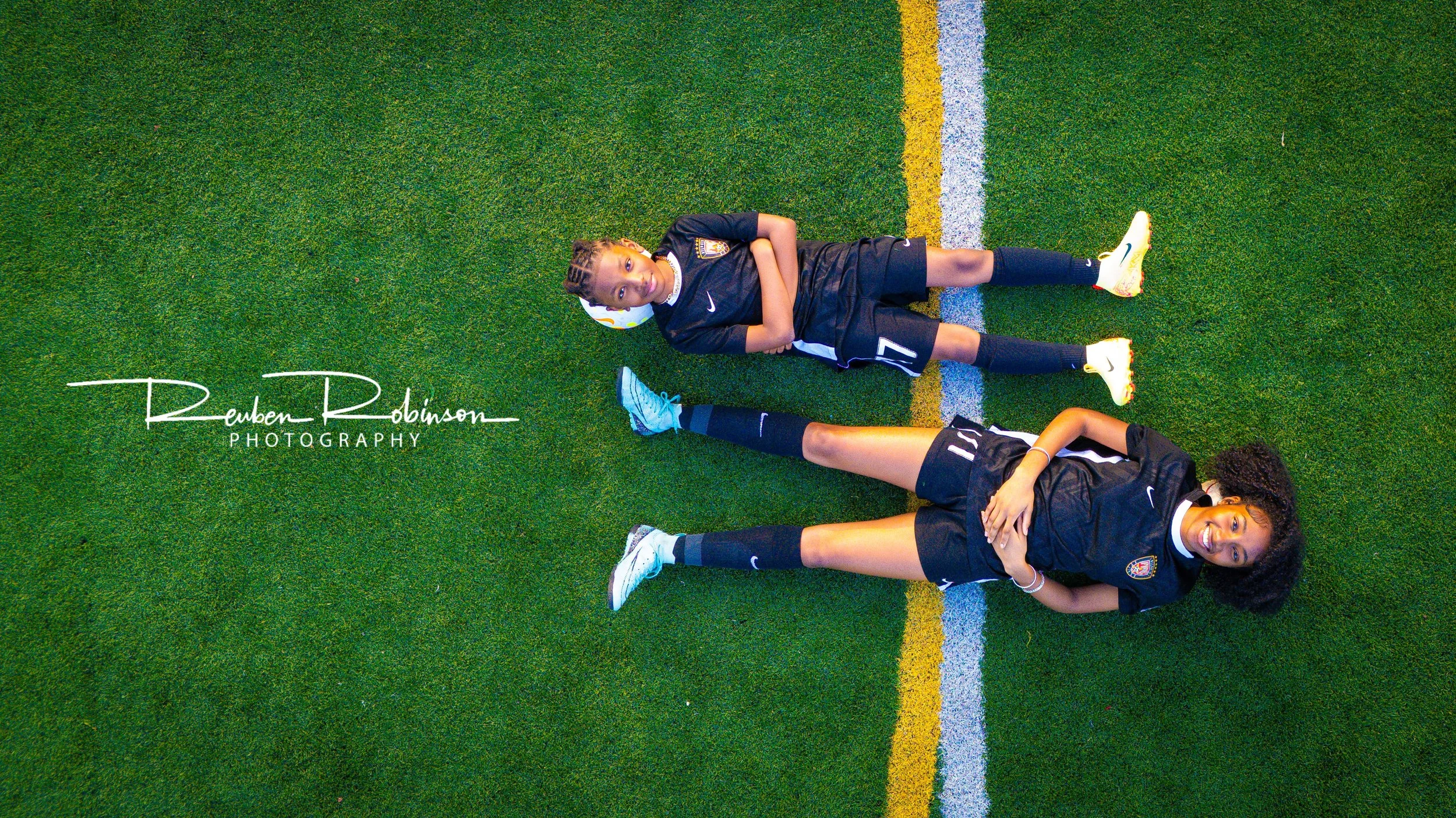 Two young female soccer players lying on a green artificial turf field, one smiling at the camera and the other partially not visible, both wearing black soccer uniforms.