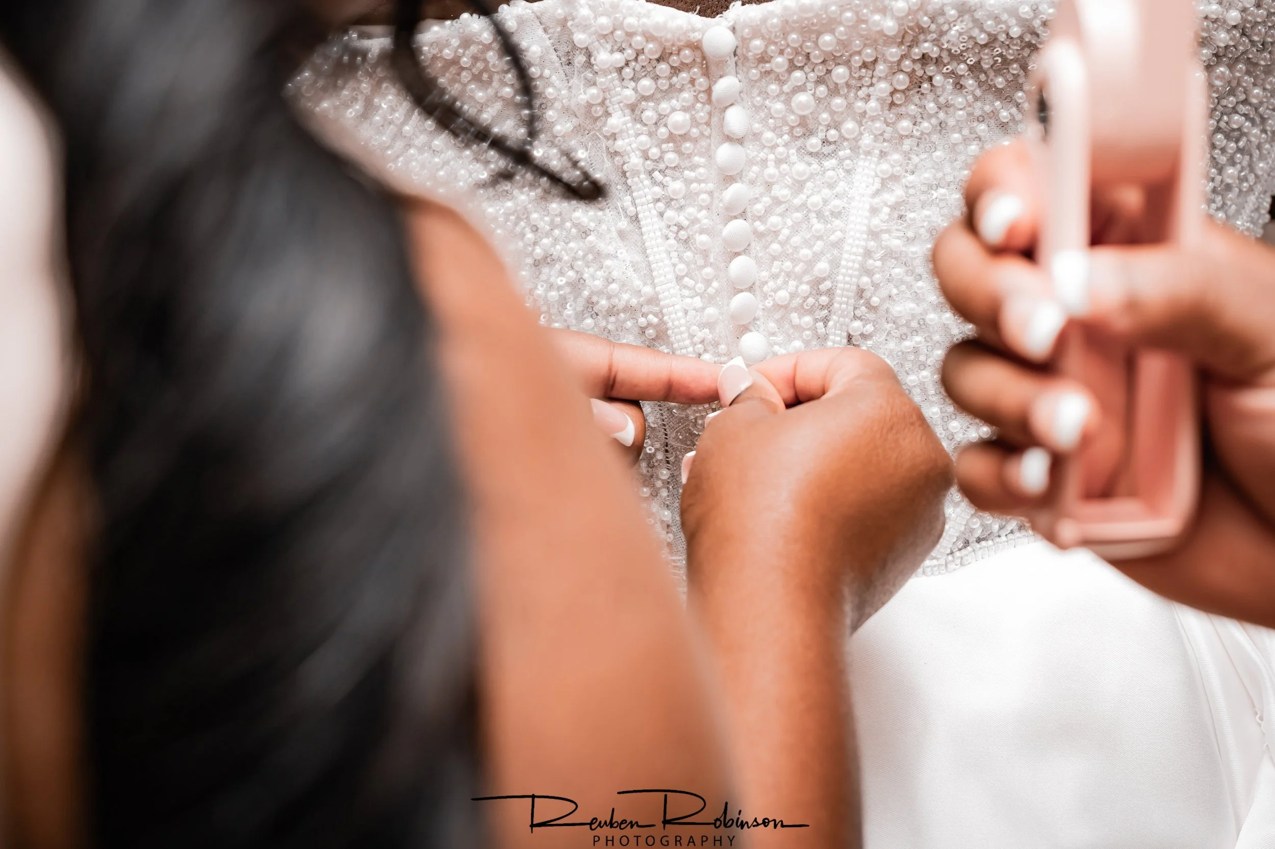 A woman is buttoning a beaded white wedding dress while another person is holding a pink mobile phone. The dress has pearl and bead embellishments.