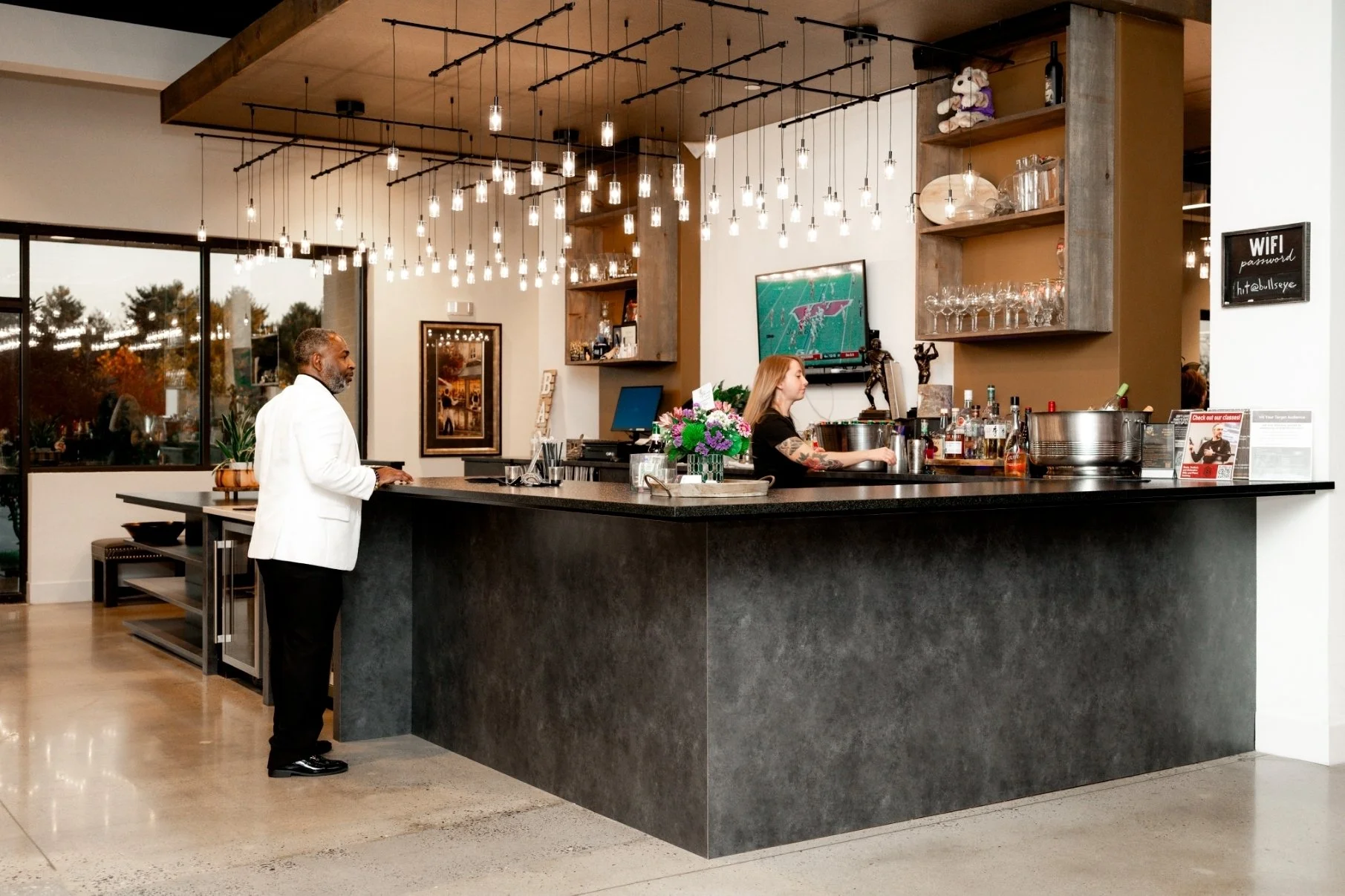 A man in a white jacket standing at a bar counter talking to a bartender in a black shirt. The bar has a dark countertop, a wall-mounted TV, and shelves with glassware and decorative items, including a teddy bear and sculptures. The area is well-lit 