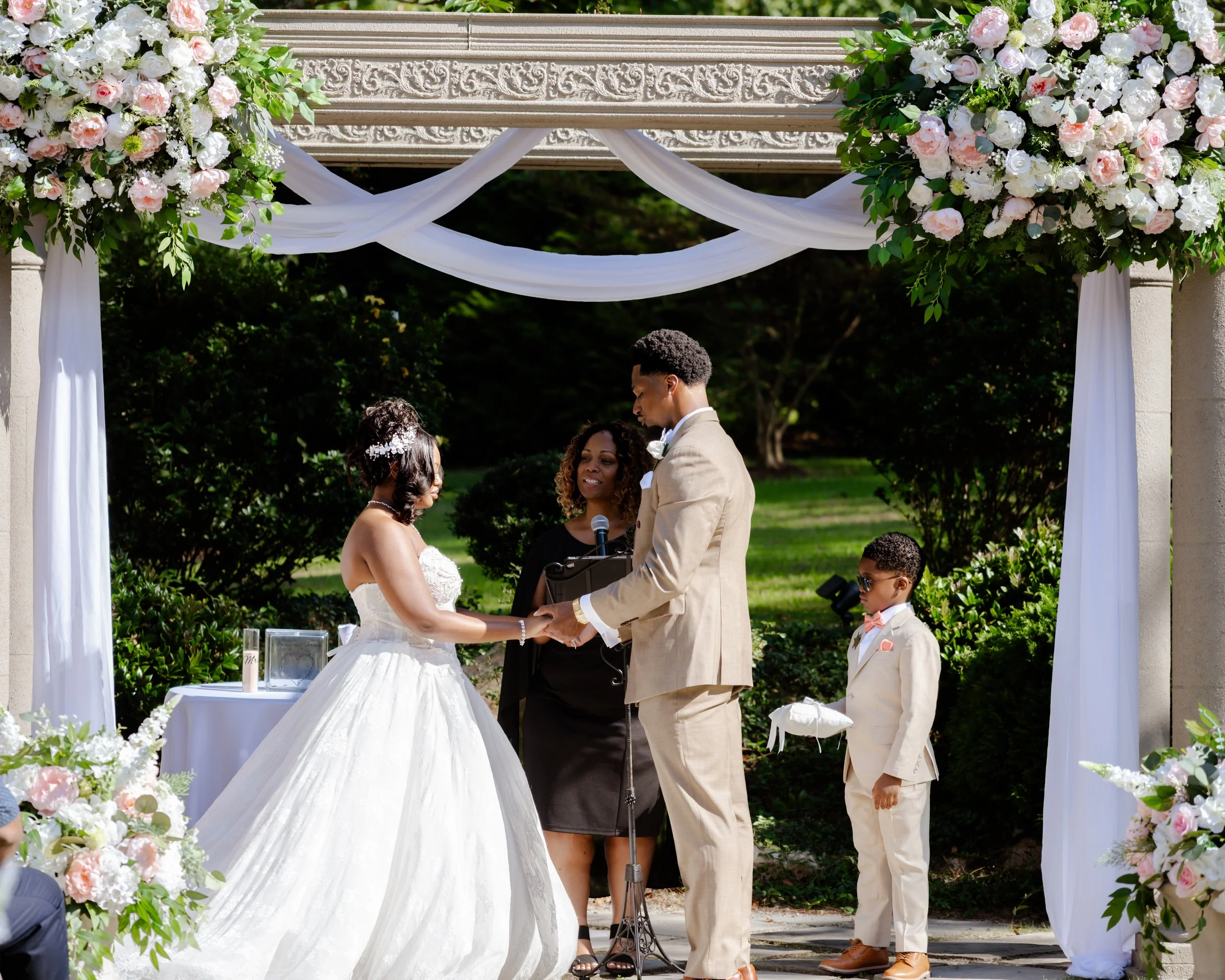 A wedding ceremony outdoors with an officiant, bride and groom holding hands under a decorated arch with pink and white flowers and white drapery, surrounded by greenery