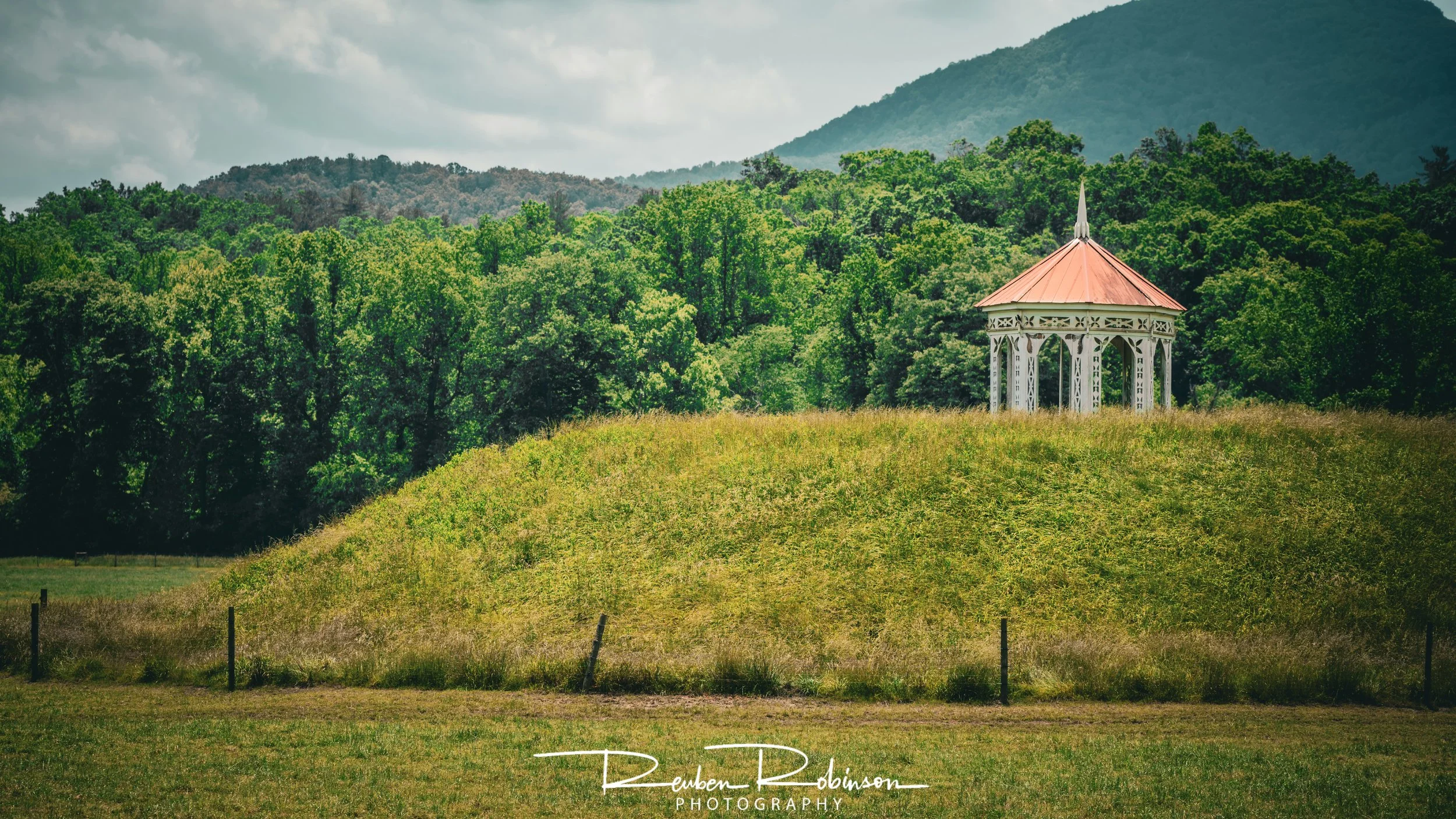 A small white gazebo with a copper-colored roof situated on a grassy hill, surrounded by dense green trees with mountains in the background.