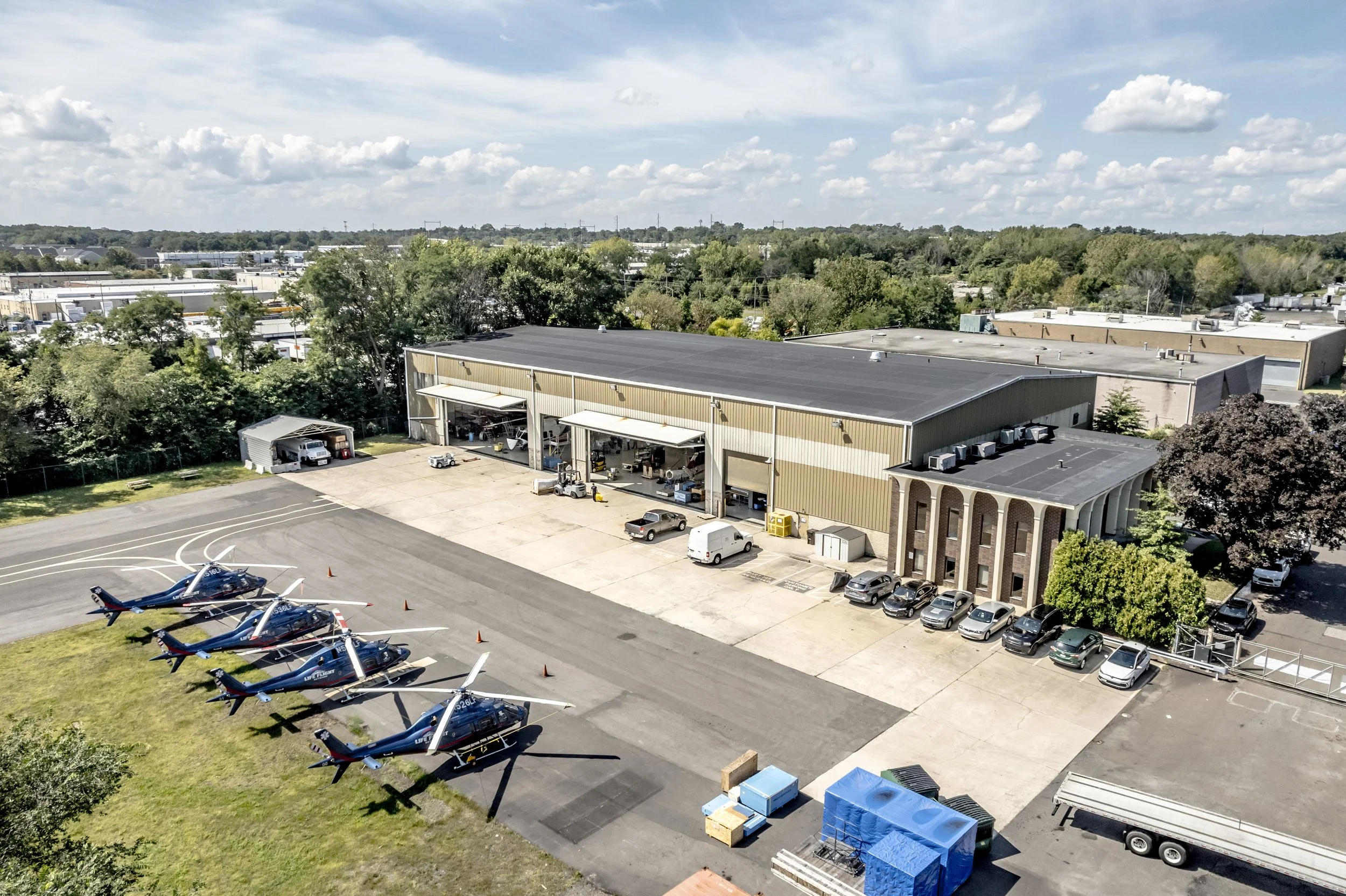 An aerial view of a helicopter hangar with five helicopters parked in front on a tarmac, surrounded by cars and trucks, with green trees and a cloudy sky in the background.