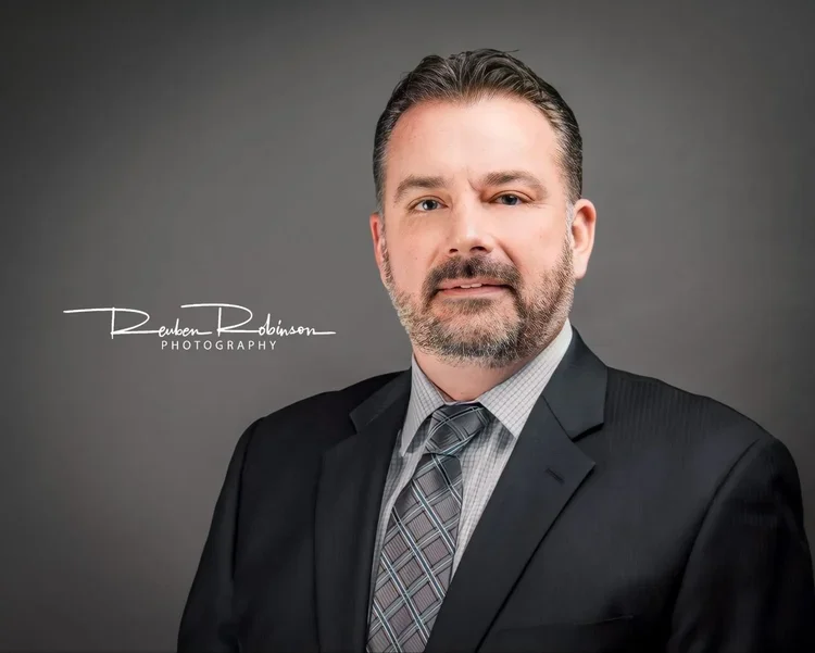 A professional headshot of a man with light skin, short dark hair, and a beard, wearing a black suit, gray checkered shirt, and patterned tie, against a gray background.