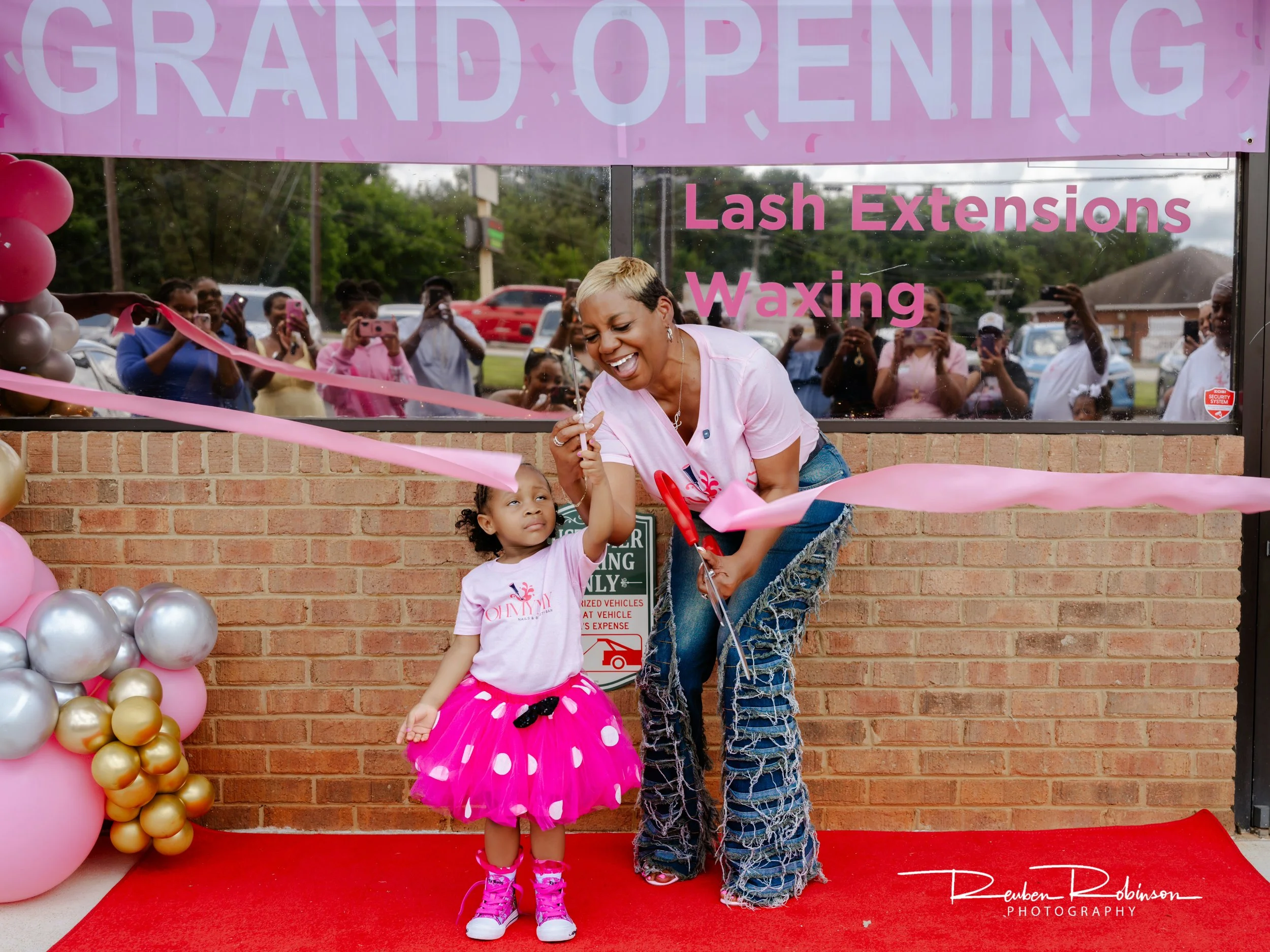 A woman and a young girl celebrate the grand opening of a salon, cutting a pink ribbon on a red carpet. The woman is smiling and the girl is looking up at her. The background includes a large pink banner with text about salon services, and a crowd of