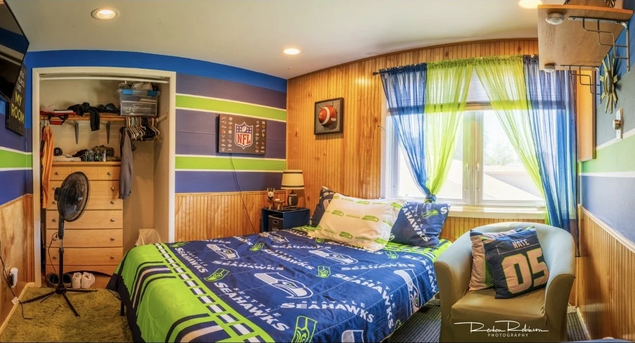 A Seahawks-themed bedroom with a bed covered in Seahawks bedding, a chair with a Seahawks pillow, a window with blue and green curtains, a small dresser, a standing fan, and NFL decor on the walls.