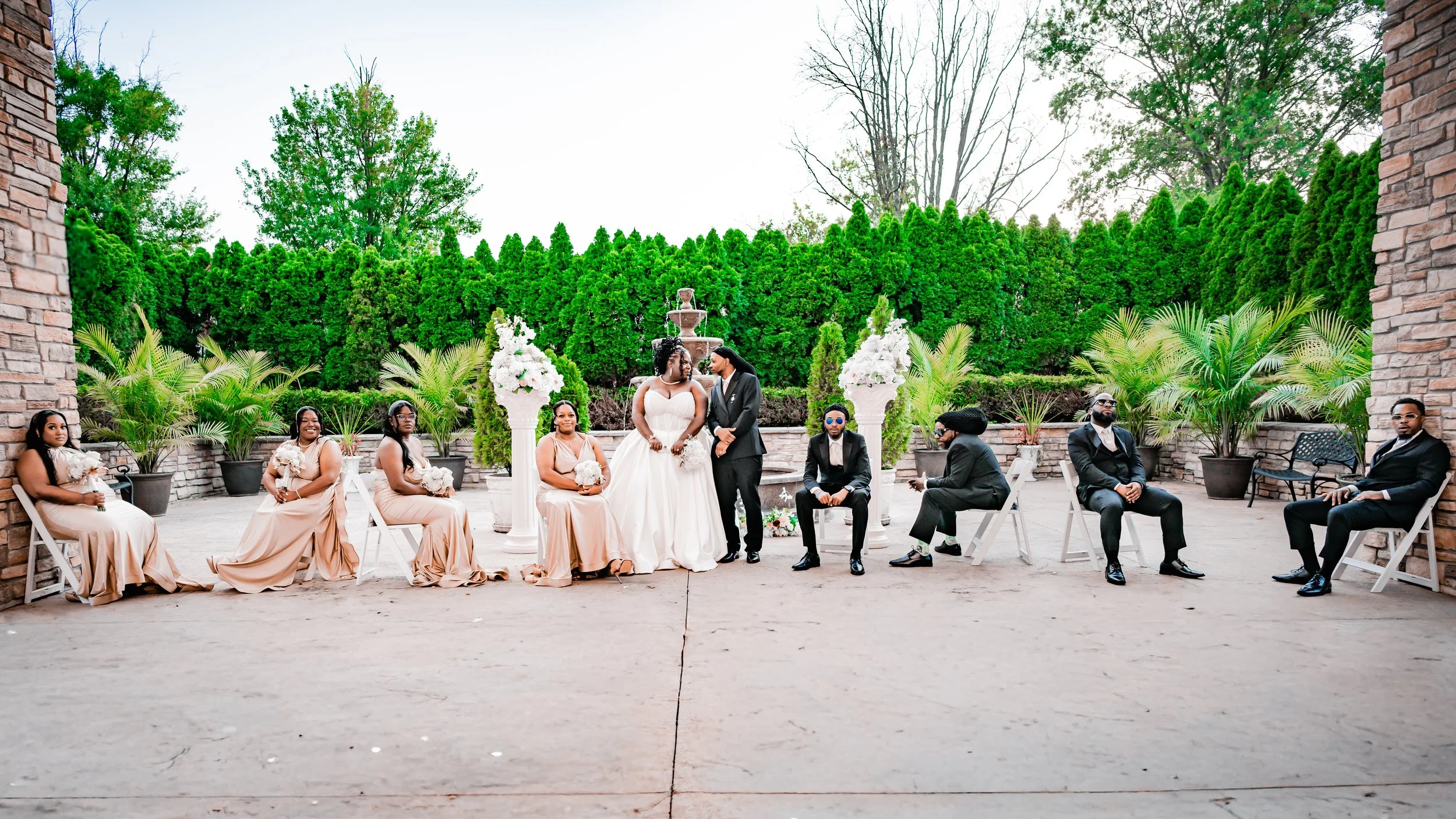 Wedding ceremony outdoors with bride and groom standing at center, surrounded by bridesmaids and groomsmen seated on white chairs, lush green trees and potted plants in background.
