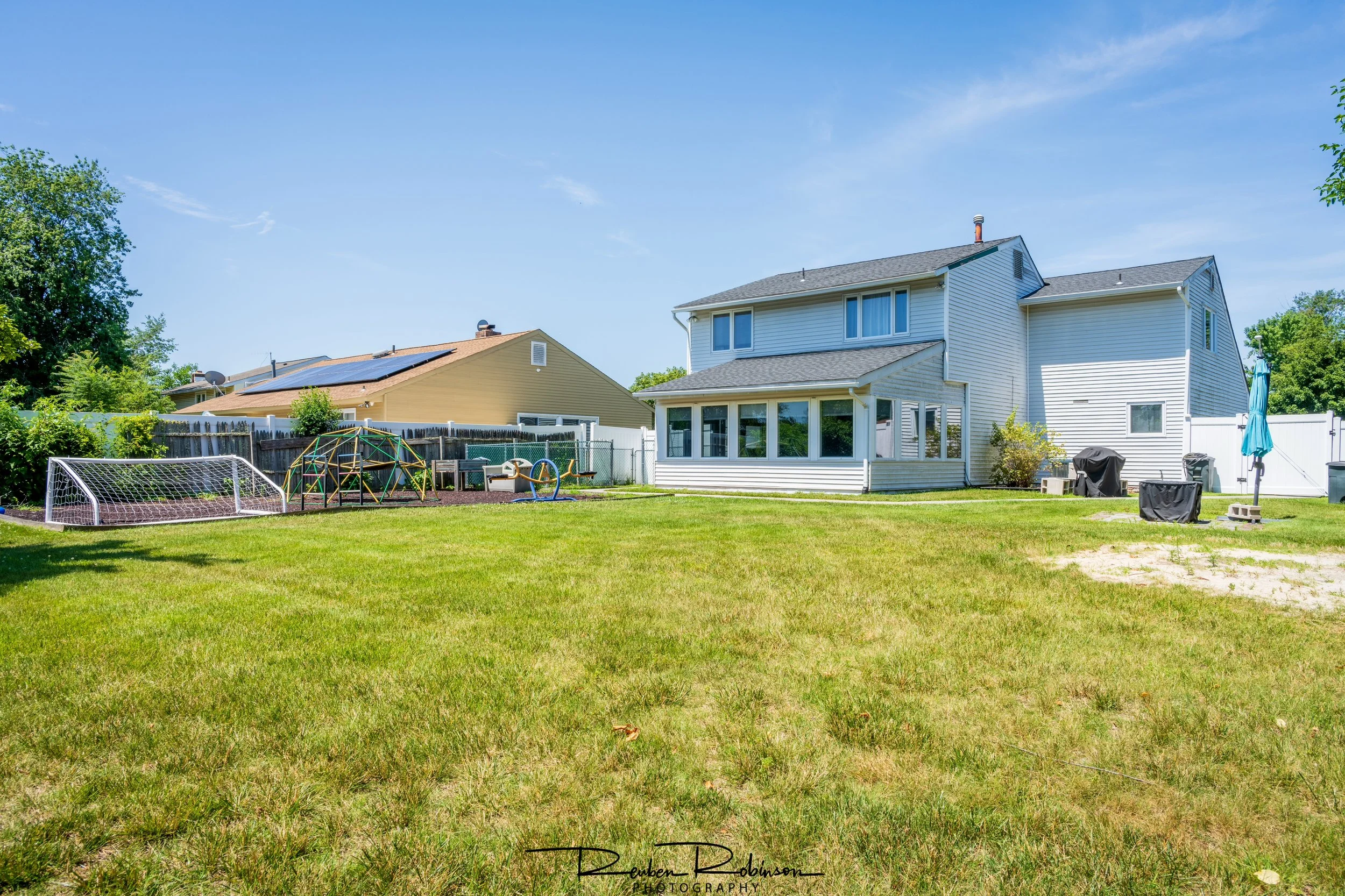 A backyard with a green lawn, a children's soccer goal, playground equipment, and a white two-story house with large windows and a small porch, under a clear blue sky with some trees and neighboring houses visible.