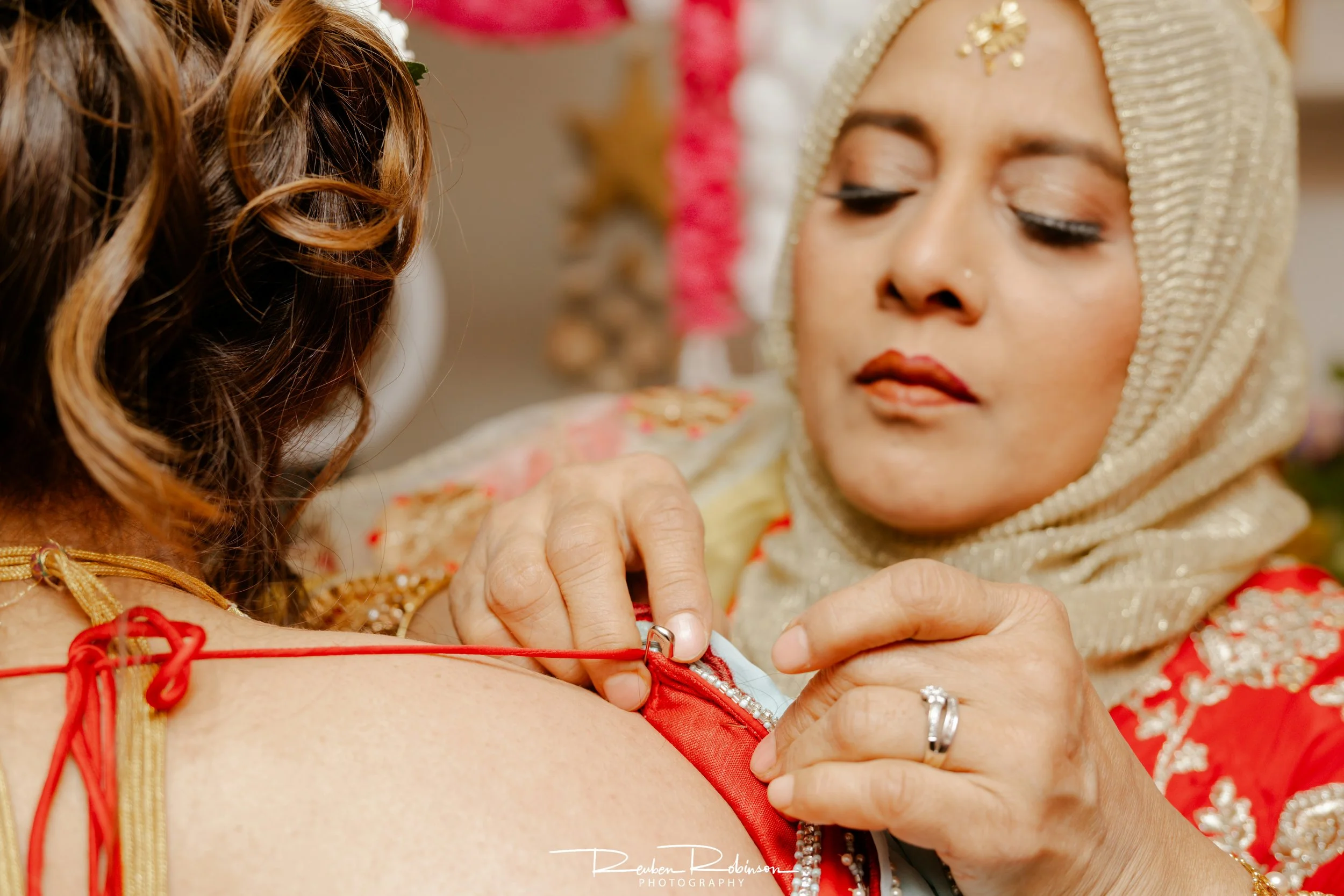 A bride in traditional wedding attire with a beige headscarf applying a red string bracelet on a woman, likely during a wedding ceremony.