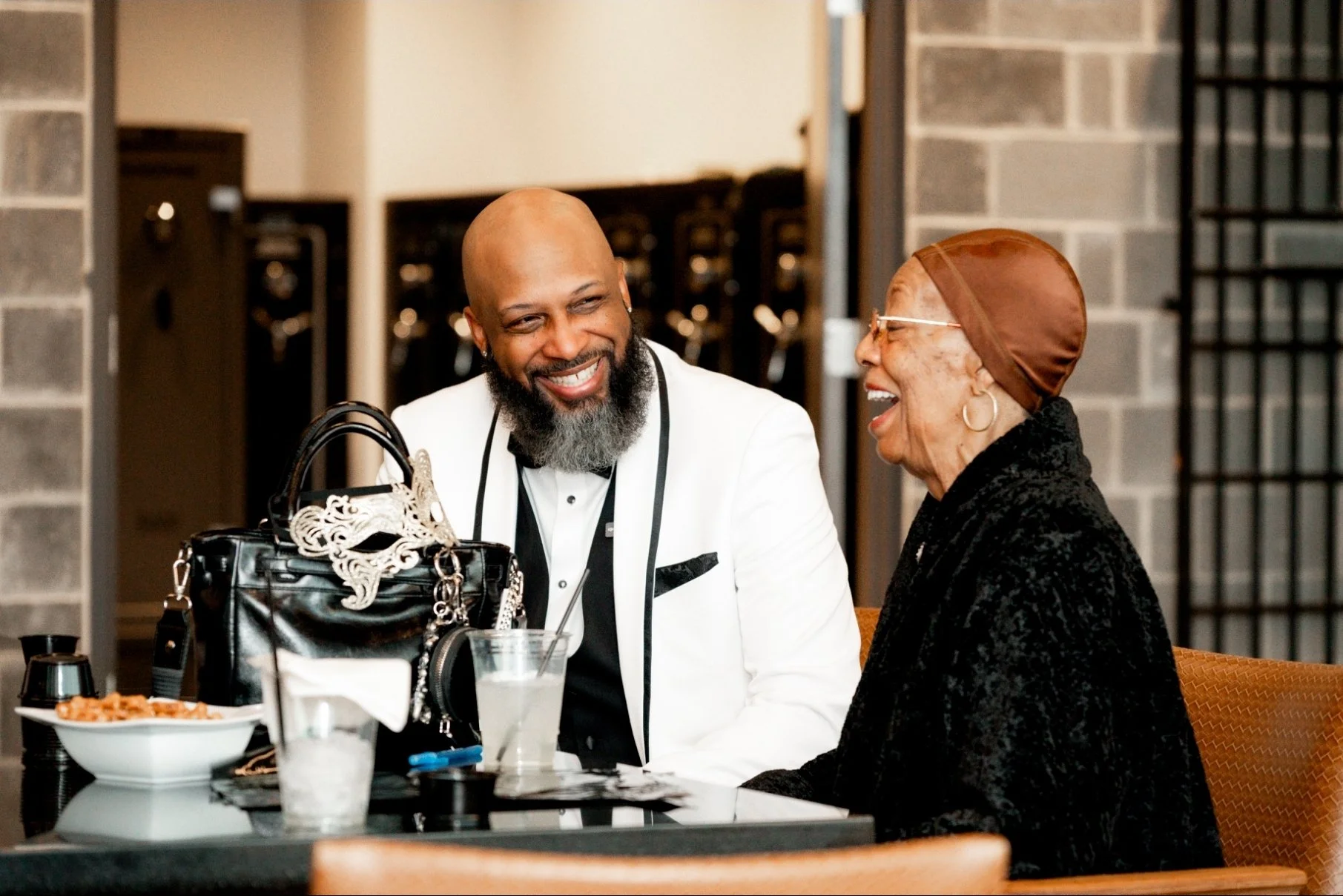 A man in a white tuxedo and a woman in a black outfit are sitting at a table, laughing and talking to each other inside a restaurant or cafe.