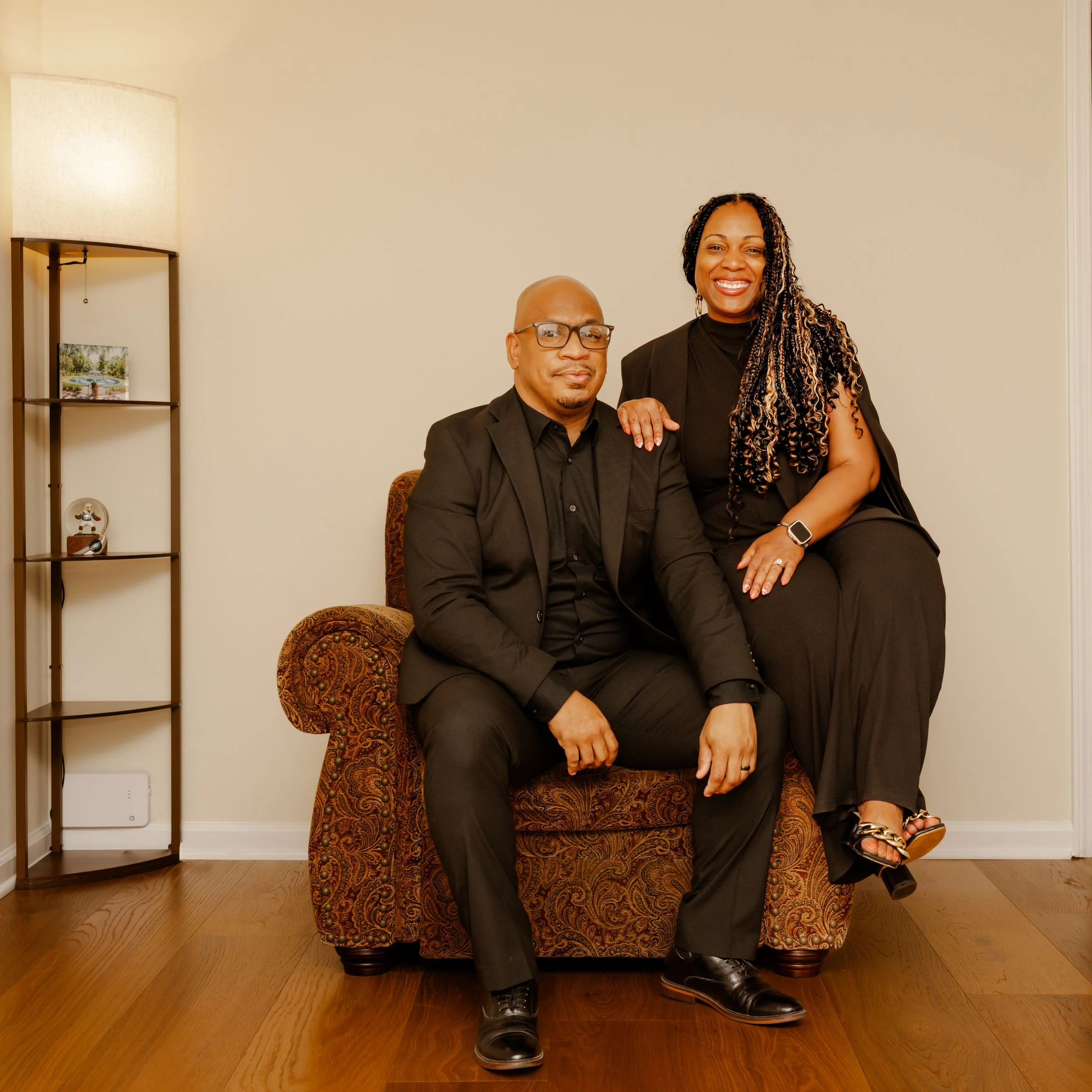 A man and a woman dressed in black sitting on a patterned armchair in a living room, smiling at the camera. The woman has long curly hair and the man is wearing glasses.