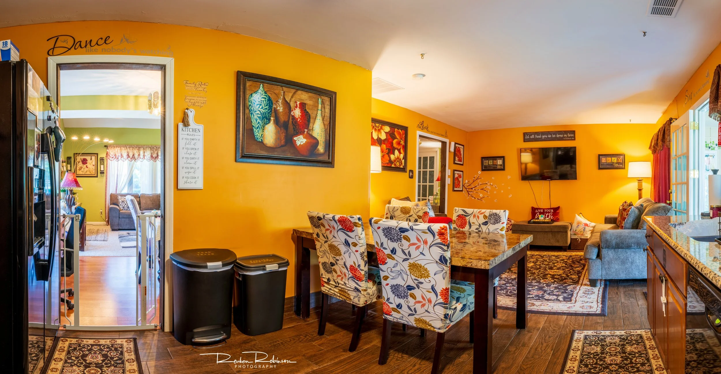 Interior view of a living room with yellow walls, a dining area with floral upholstered chairs, a gray sofa, wall decorations, and a mounted flat-screen TV. There's a sliding door to an outdoor patio.
