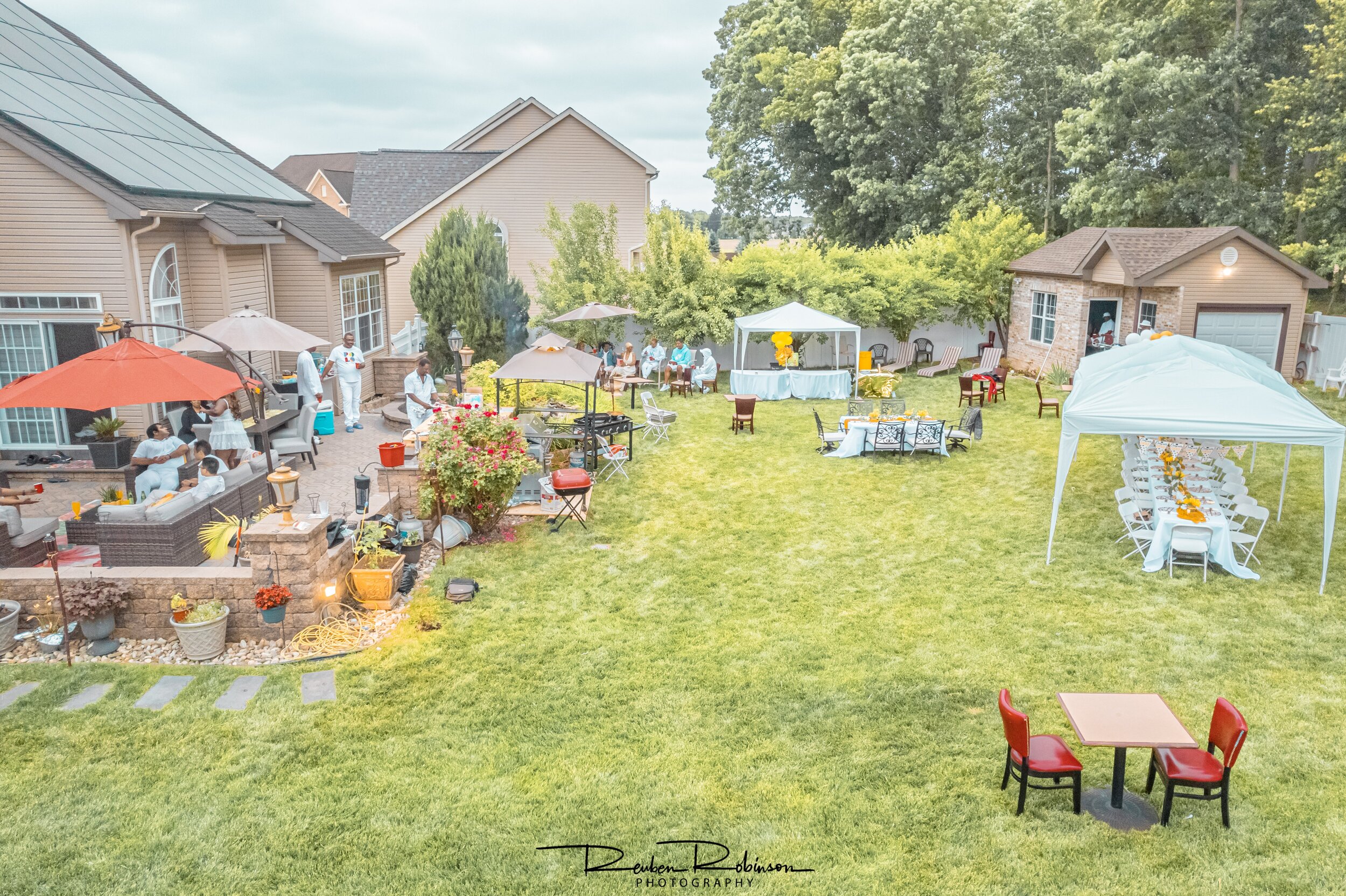 Backyard event with people, tents, tables, and chairs, featuring a patio area, umbrellas, and a garden, with a house and trees in the background.