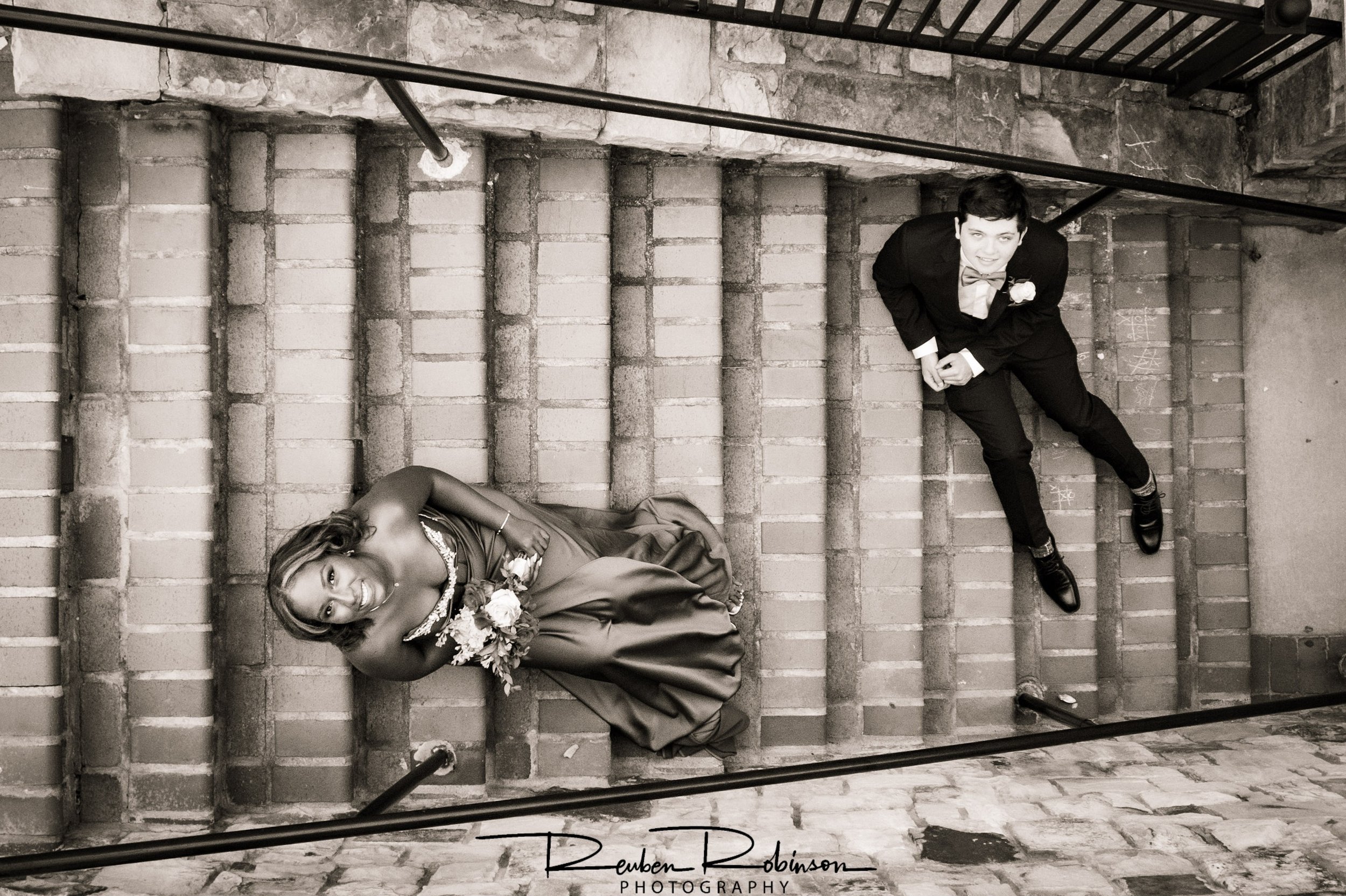 Black and white photo of a woman lying on a staircase in a gown holding a bouquet, and a man in a suit standing above her, both on brick steps.