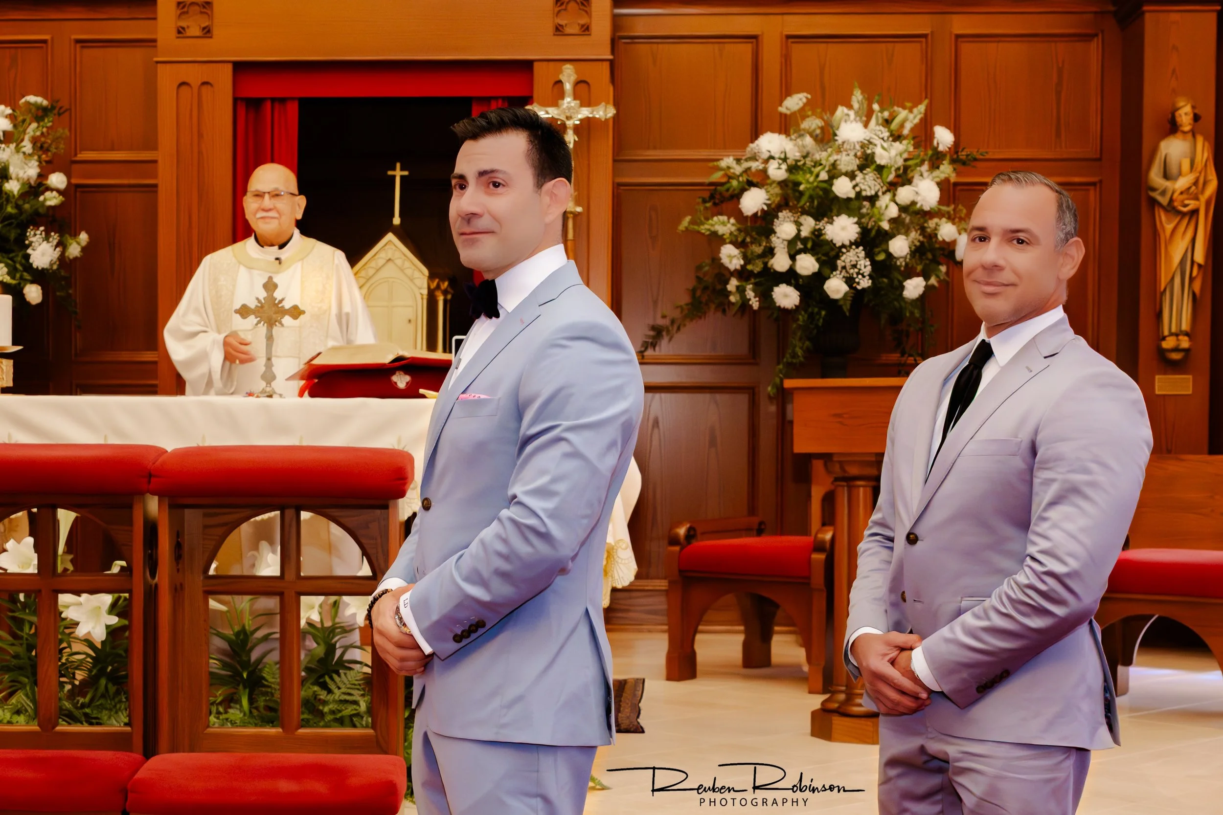A groom and another man stand in a church during a wedding ceremony, with the altar and floral arrangements visible in the background.