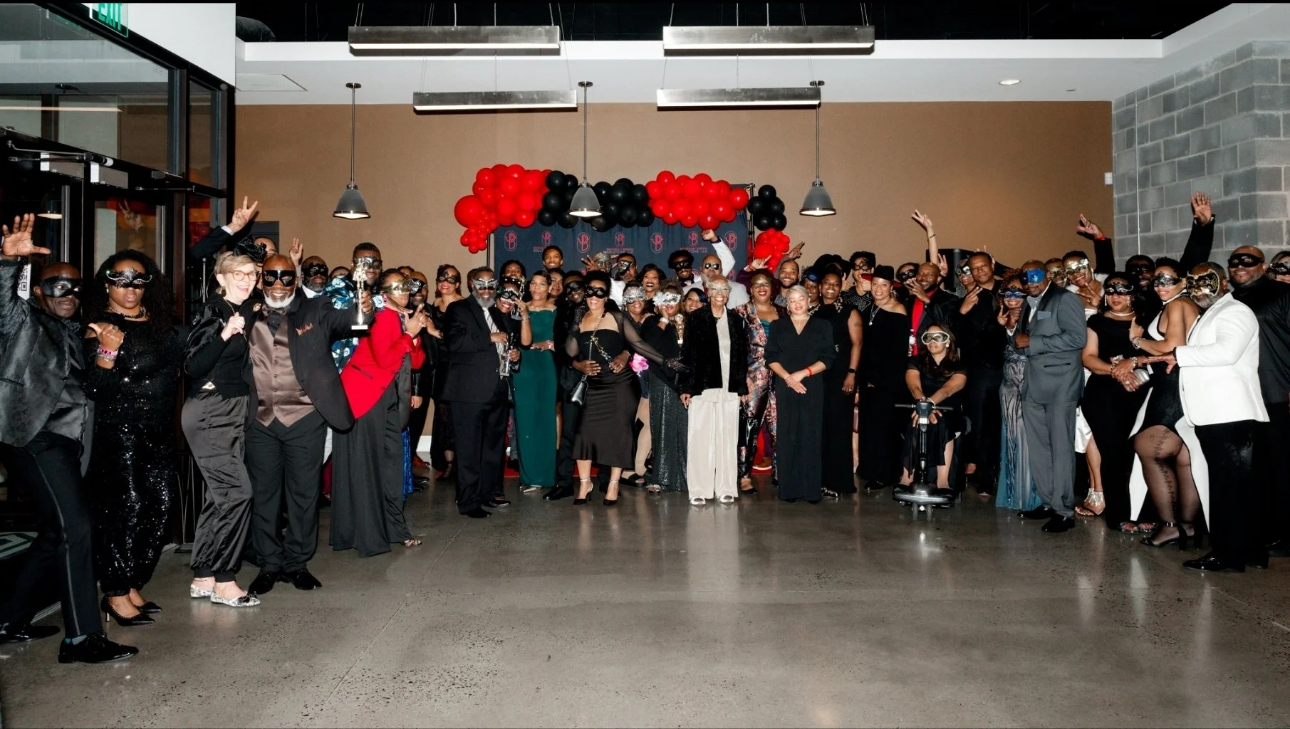 Group of people dressed formally for a celebration or event, wearing party masks, standing in front of a backdrop with red and black balloons, inside a modern venue.
