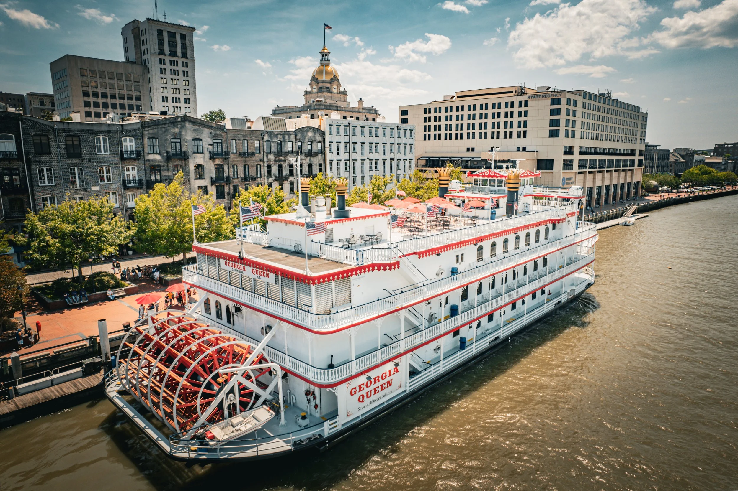 A riverboat named Georgia Queen docked at a pier along a city waterfront with trees, shops, and tall buildings in the background on a partly cloudy day.
