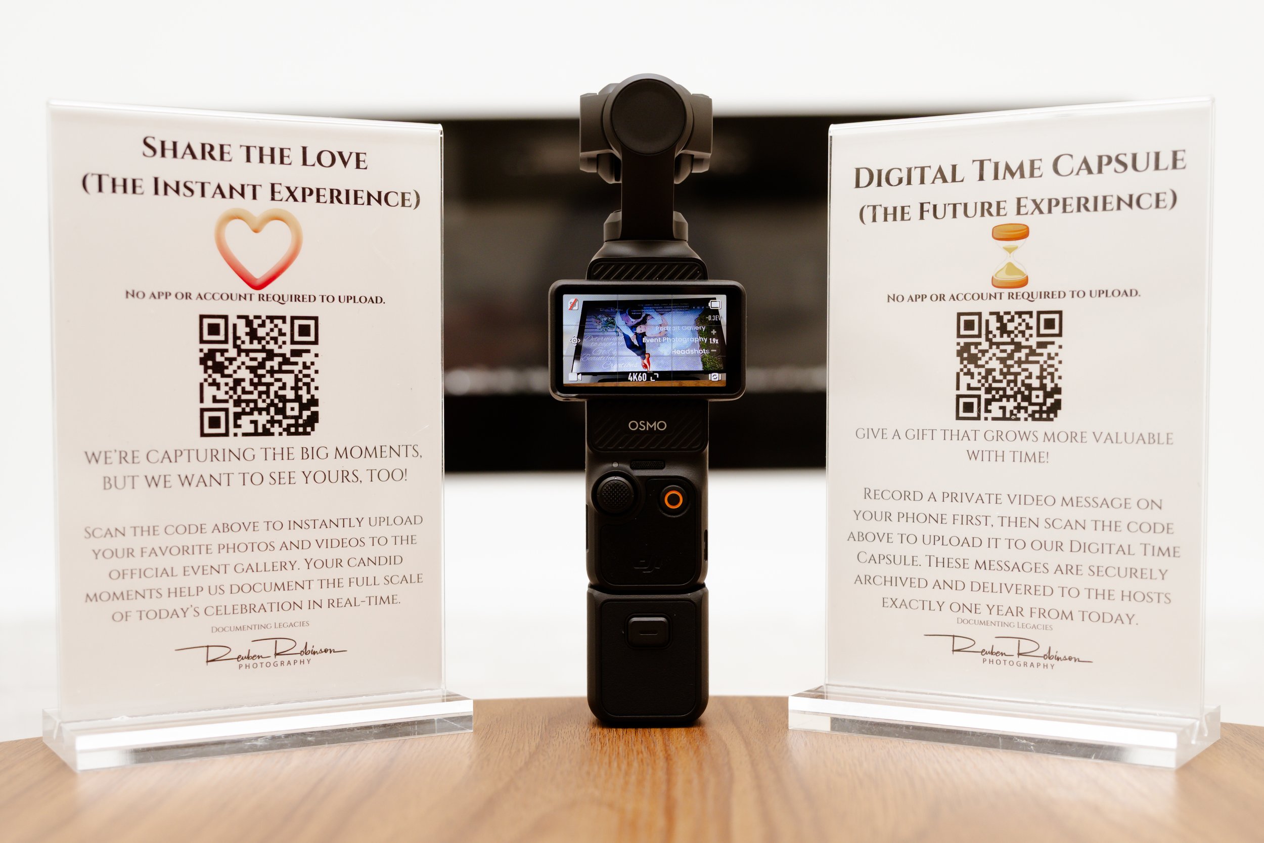 A digital camera on a stand with two informational signs on either side. The signs mention sharing a love story through photos and creating a digital time capsule, both with QR codes.