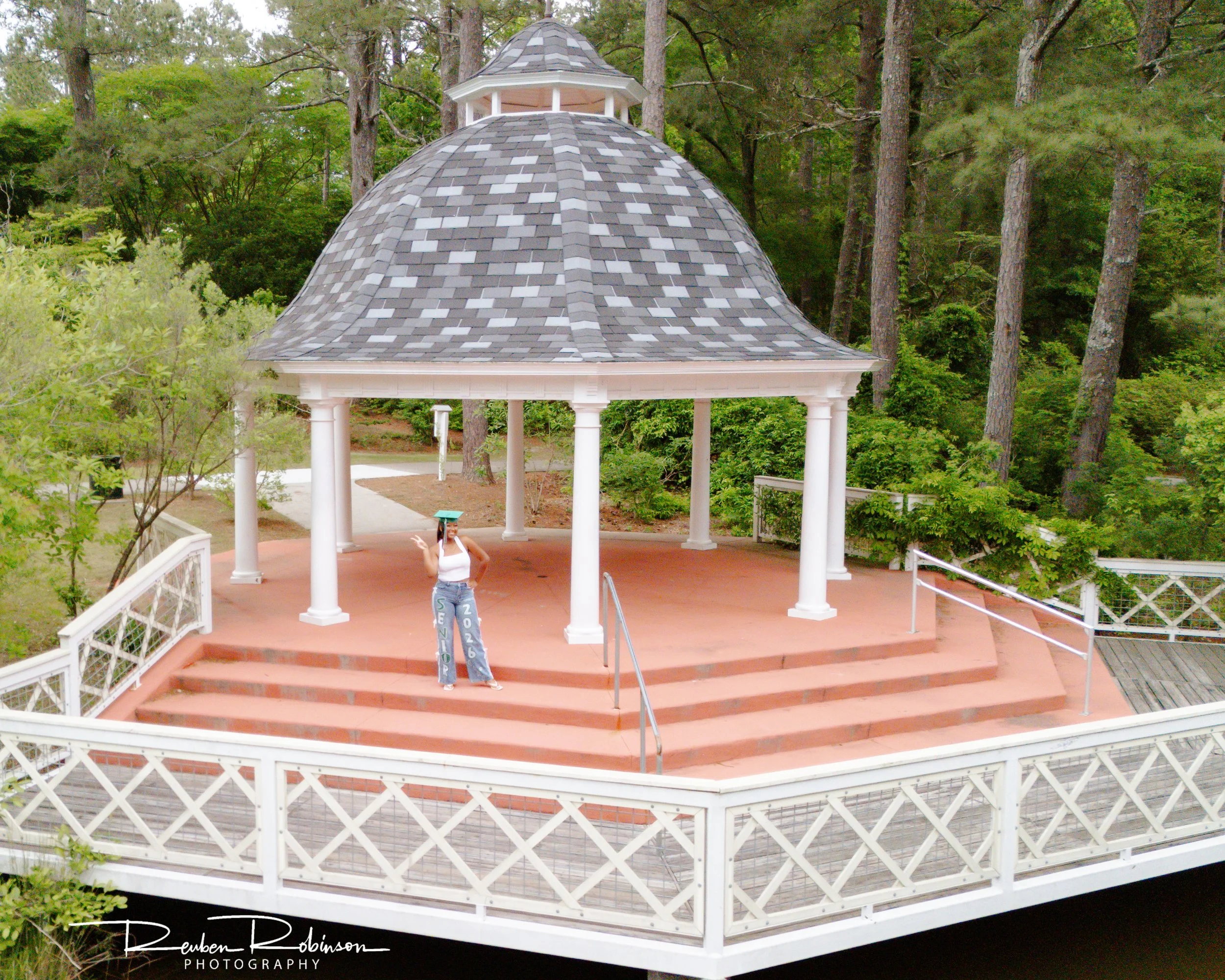 A person standing on the steps of a white bandstand with a shingled, gray lid and surrounded by trees in a park.