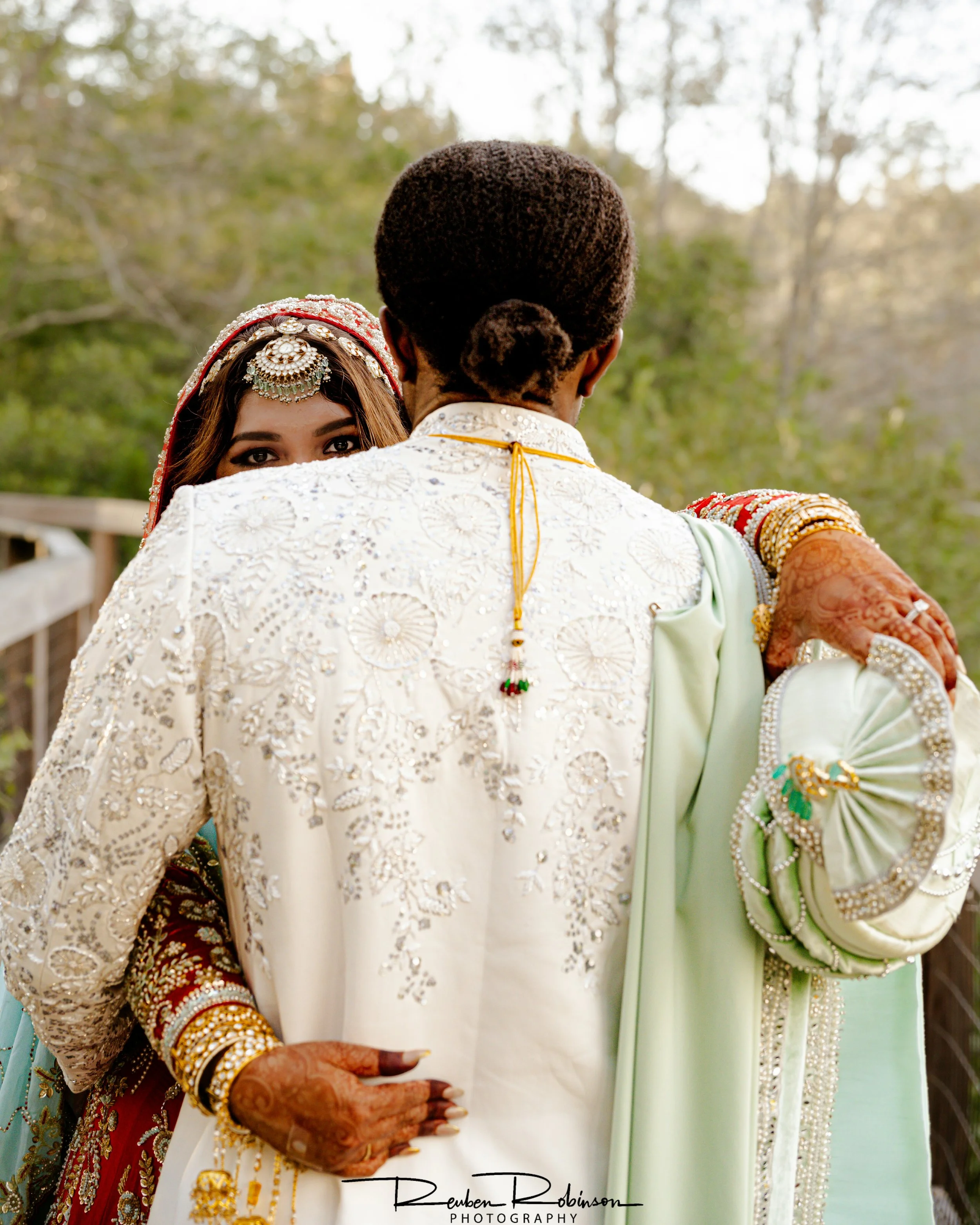 A bride and groom are standing outdoors, seen from behind. The bride is wearing traditional Indian attire with jewelry, and the groom is in a light-colored embroidered outfit. The bride's eyes are visible over the groom's shoulder, and they are embra