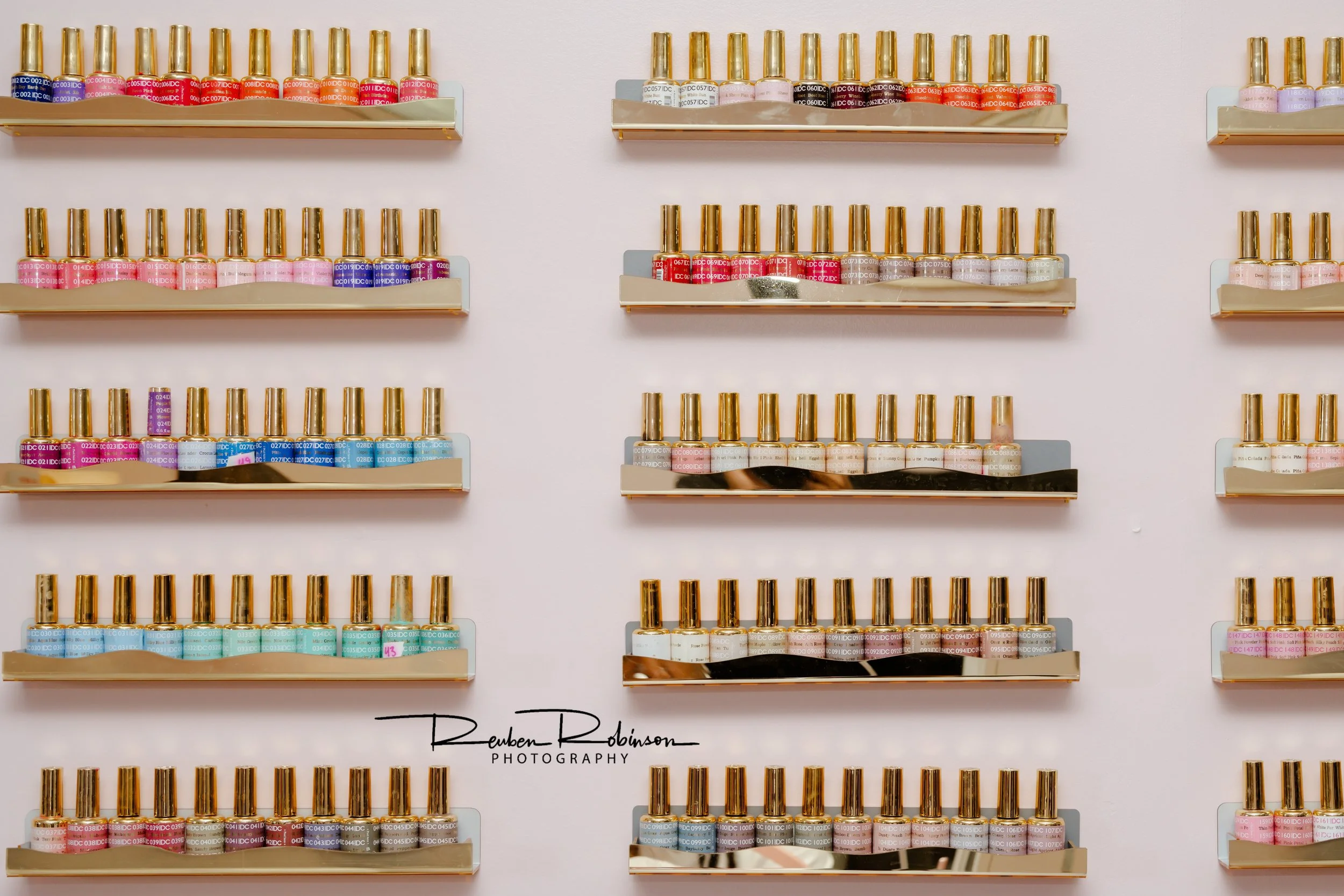 Display of multiple rows of small bottles with golden caps on beige shelves, containing various colors of liquid nail polish, arranged on a light pink wall.