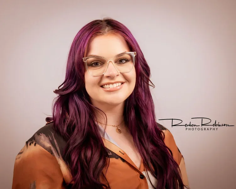 A young woman with long purple hair and glasses, smiling at the camera, wearing a brown and black shirt, and a necklace, against a plain background.