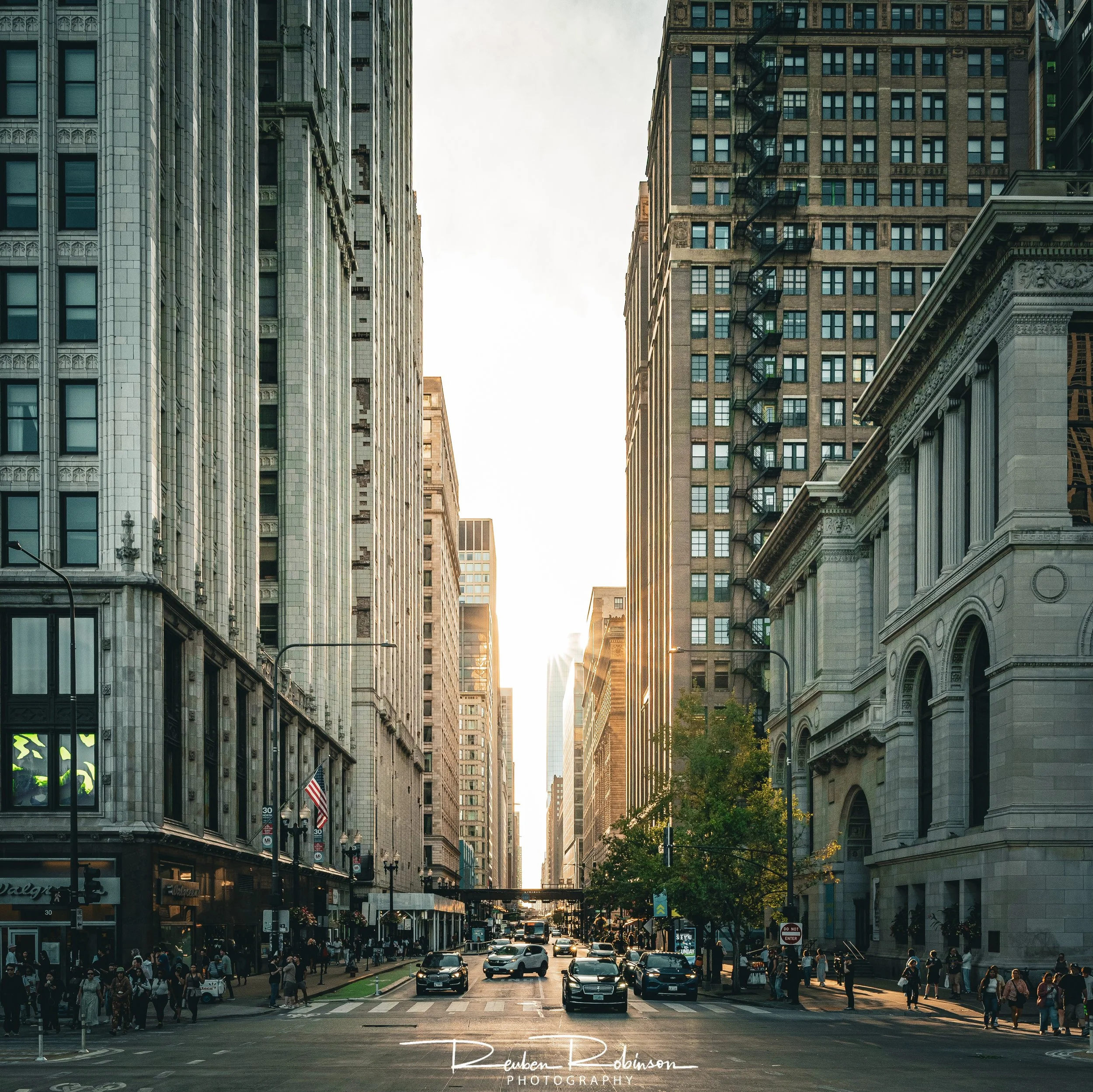Busy city street with people walking and cars driving, tall buildings lining both sides, sunlight shining between the buildings.