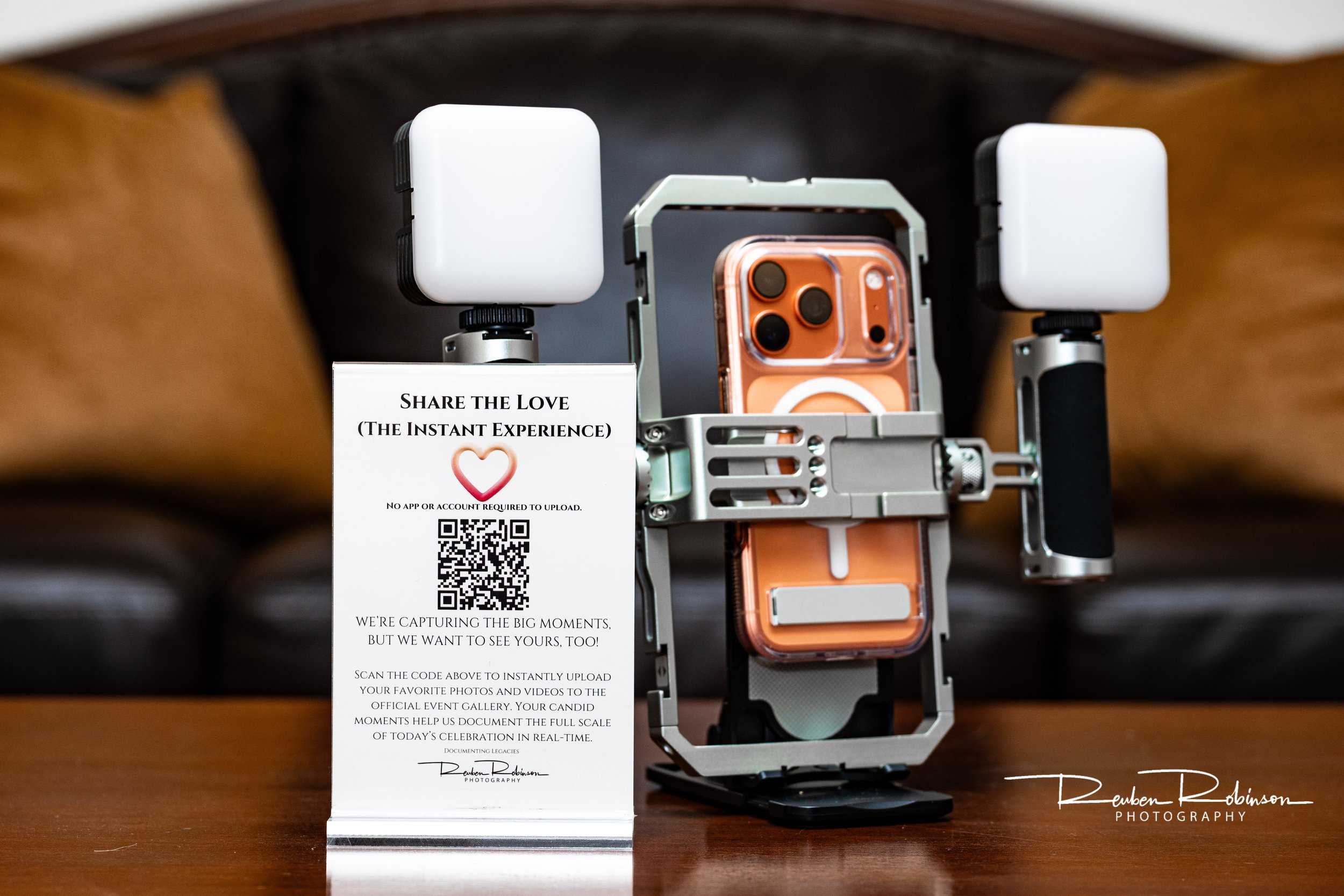 A camera setup with two white lights, a smartphone in a robotic mount, and a sign about sharing moments through photos and videos, with a QR code for instant uploads.