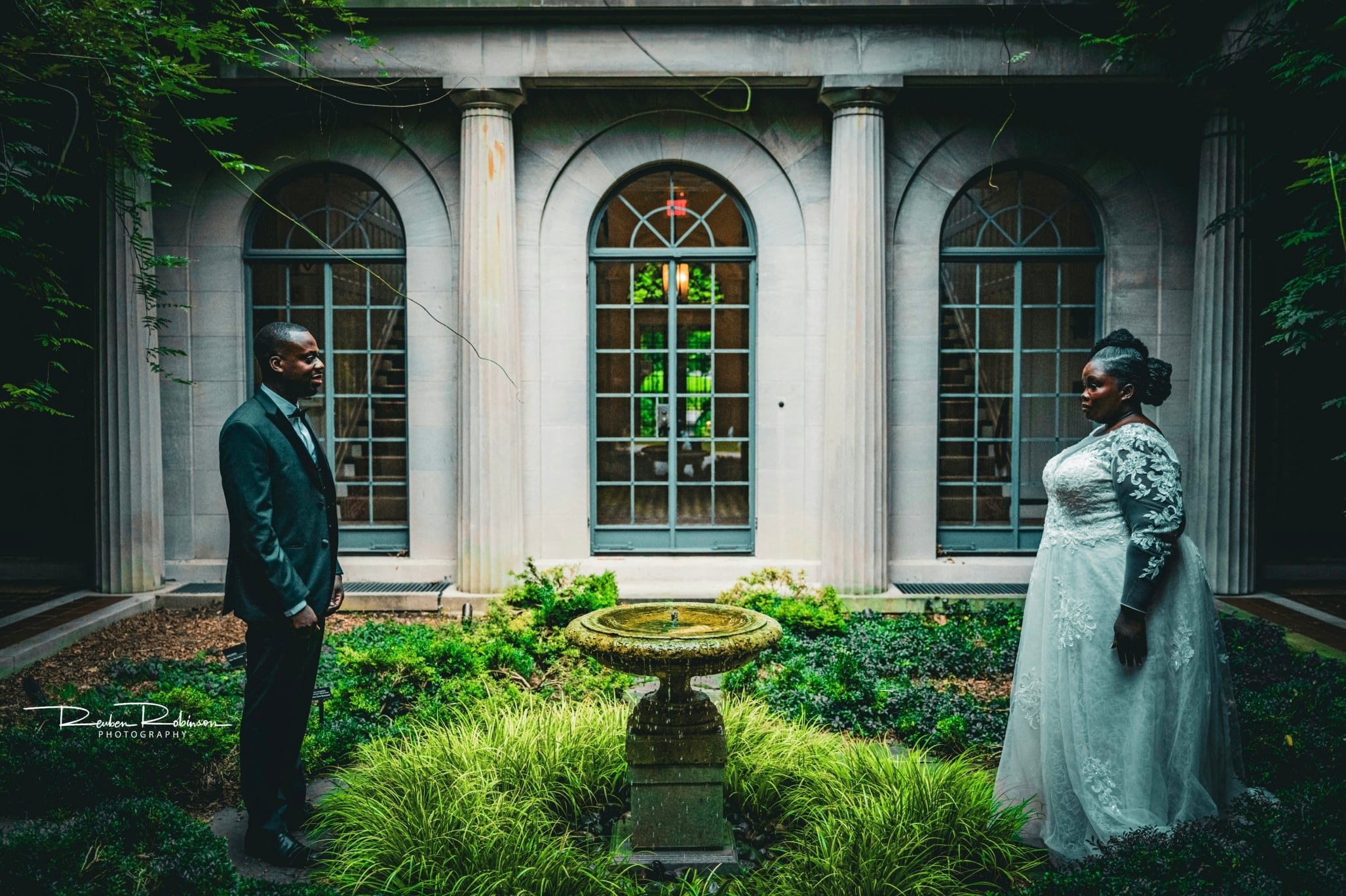 A wedding couple faces each other in an elegant garden courtyard, with classical architecture in the background and a decorative fountain in the foreground.