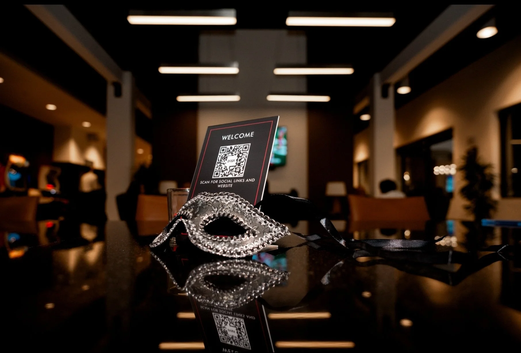 A decorative silver bracelet with rhinestones, a black face mask, and a card with a QR code on a shiny black surface, in a dimly lit indoor setting.