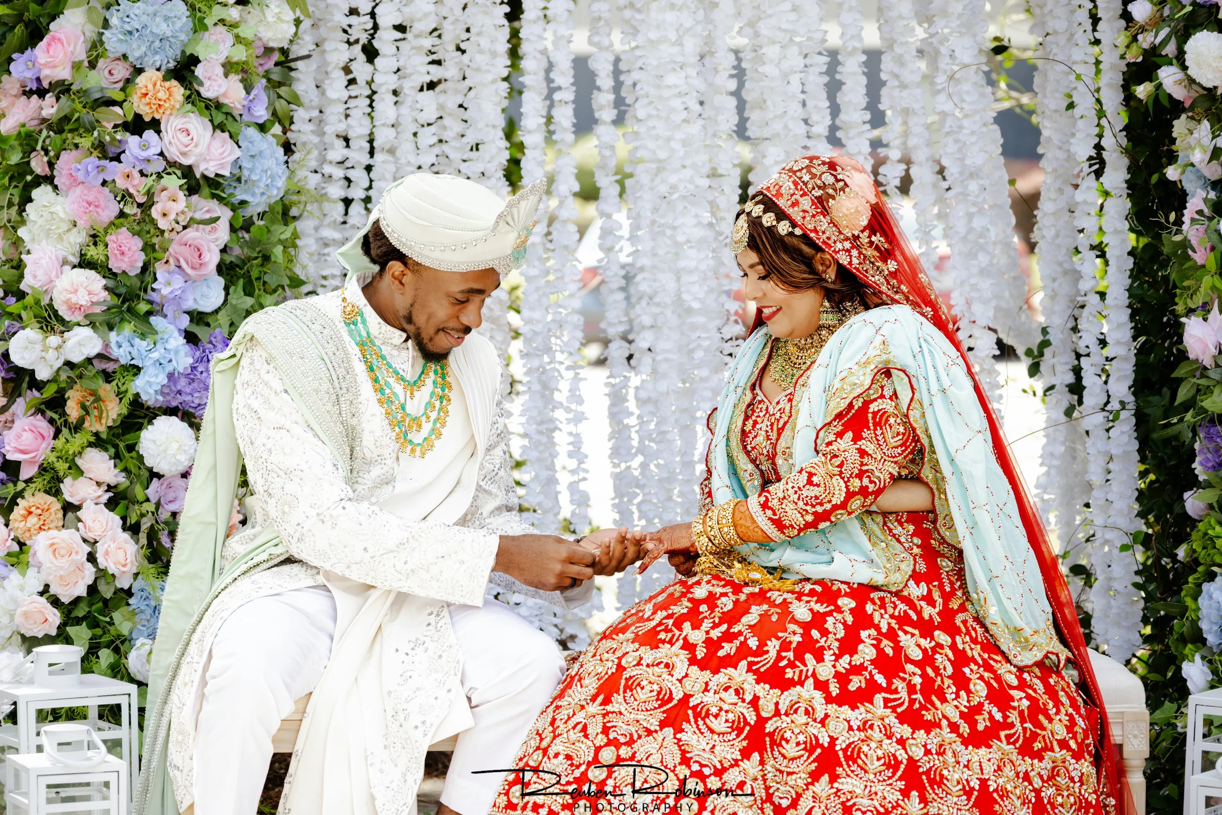 A bride and groom sit together during a wedding ceremony, surrounded by floral decorations and white hanging flowers. The groom is dressed in white traditional attire with a turban, and the bride wears a red and gold bridal dress with intricate embro