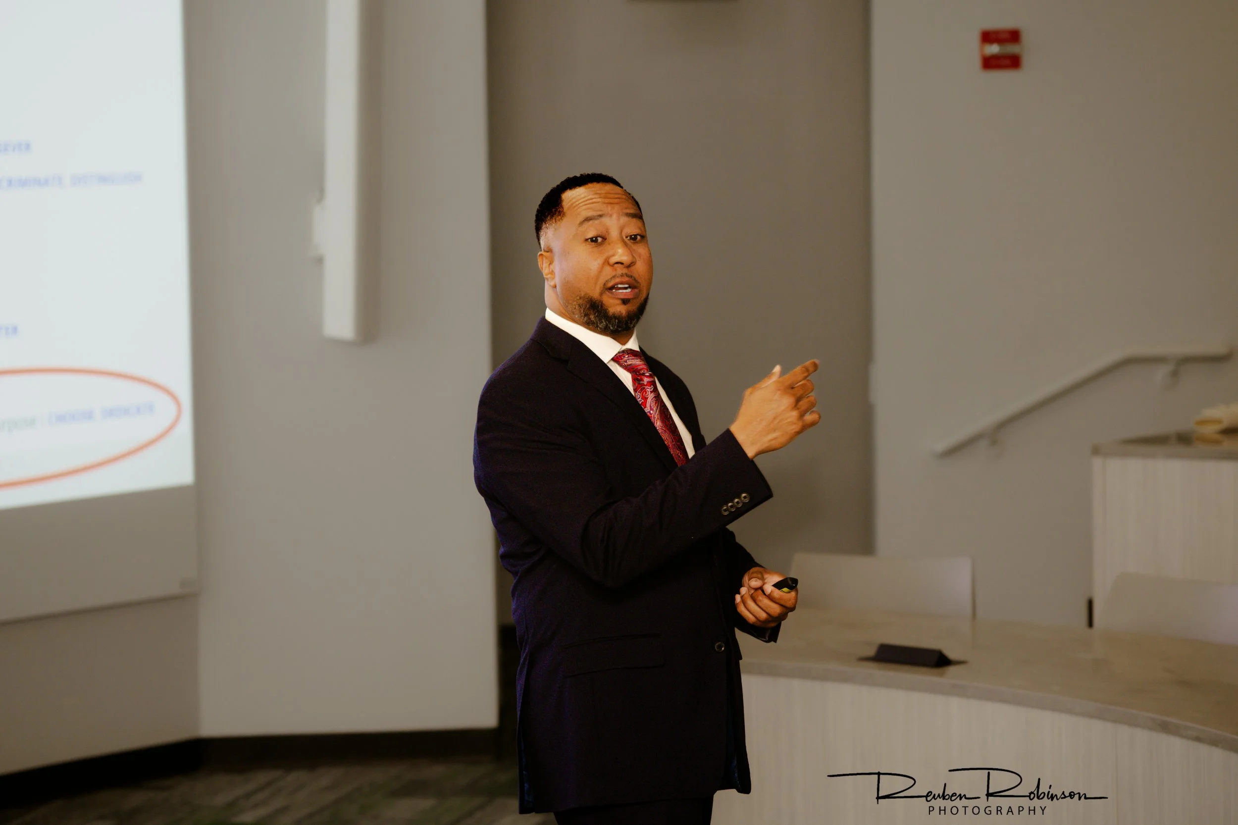 Man in a business suit giving a presentation, pointing at a whiteboard on the wall in a room.