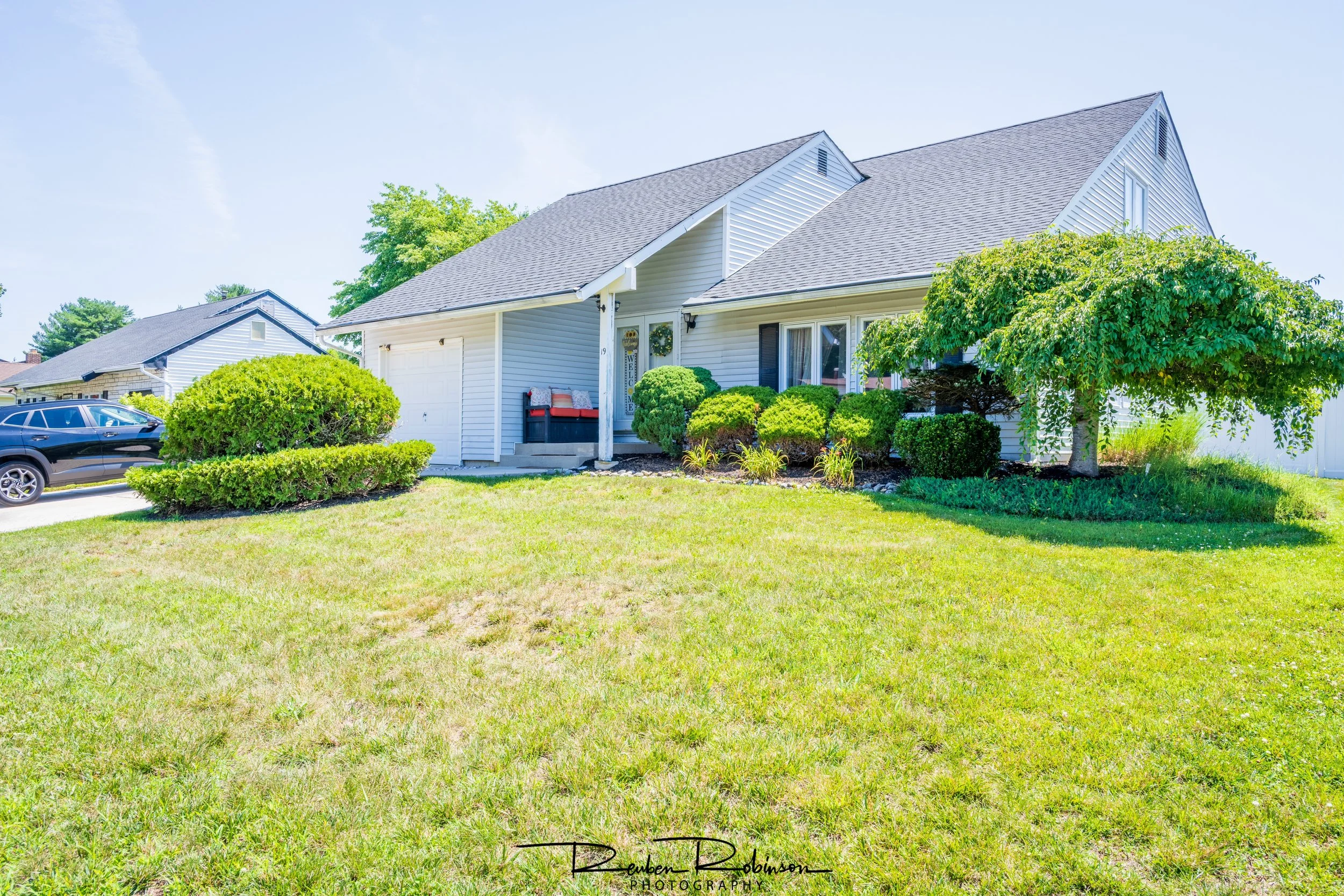 A white house with a gray roof and black shutters, surrounded by green bushes and trees, with a black car parked in the driveway under a blue sky.