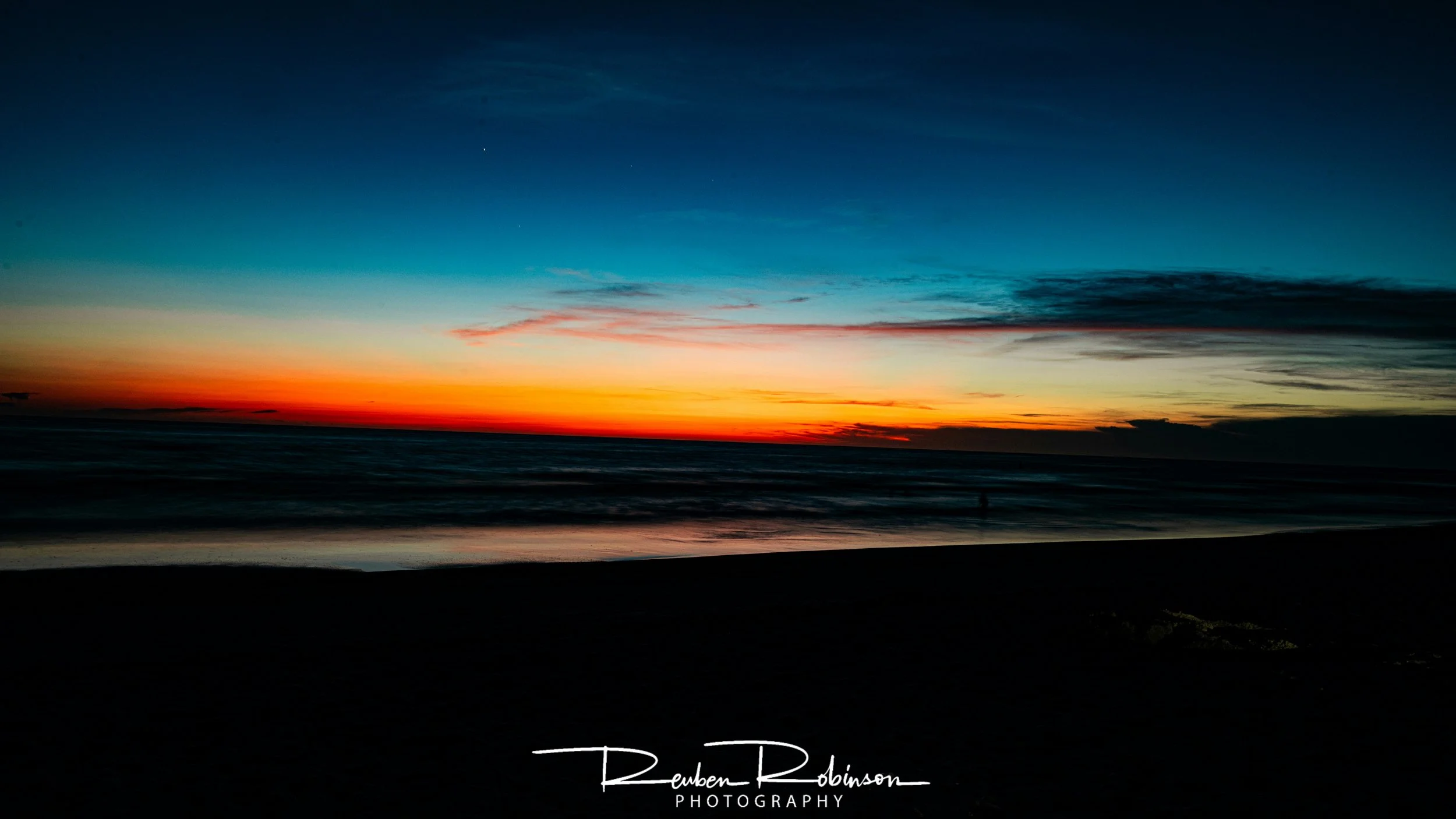 A colorful sunset over the ocean with a dark silhouette of the shoreline in the foreground and the sky transitioning from orange and yellow near the horizon to deep blue at the top.