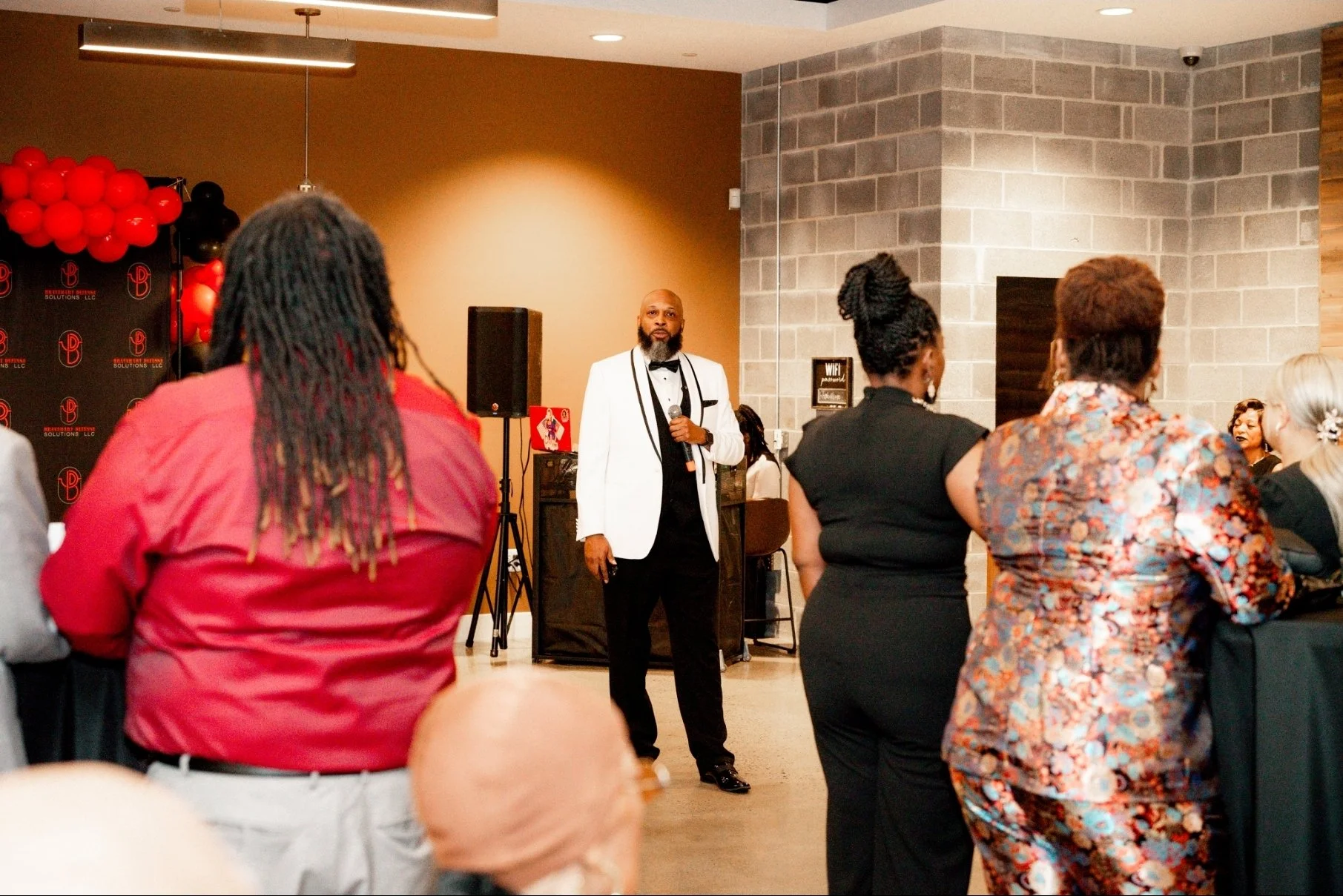 Man in a white tuxedo jacket, black bow tie, and black pants standing with a microphone in hand, speaking to an audience at an indoor event with a decorated backdrop and various seated guests.